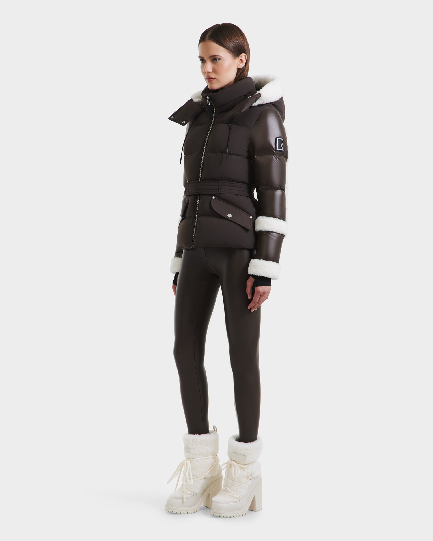 A woman wearing the Rudsak AUDREY - 8124582 Espresso, a fitted dark brown puffer jacket with white shearling trim and RDS certified down, pairs it with matching leggings and white platform boots against a white background.
