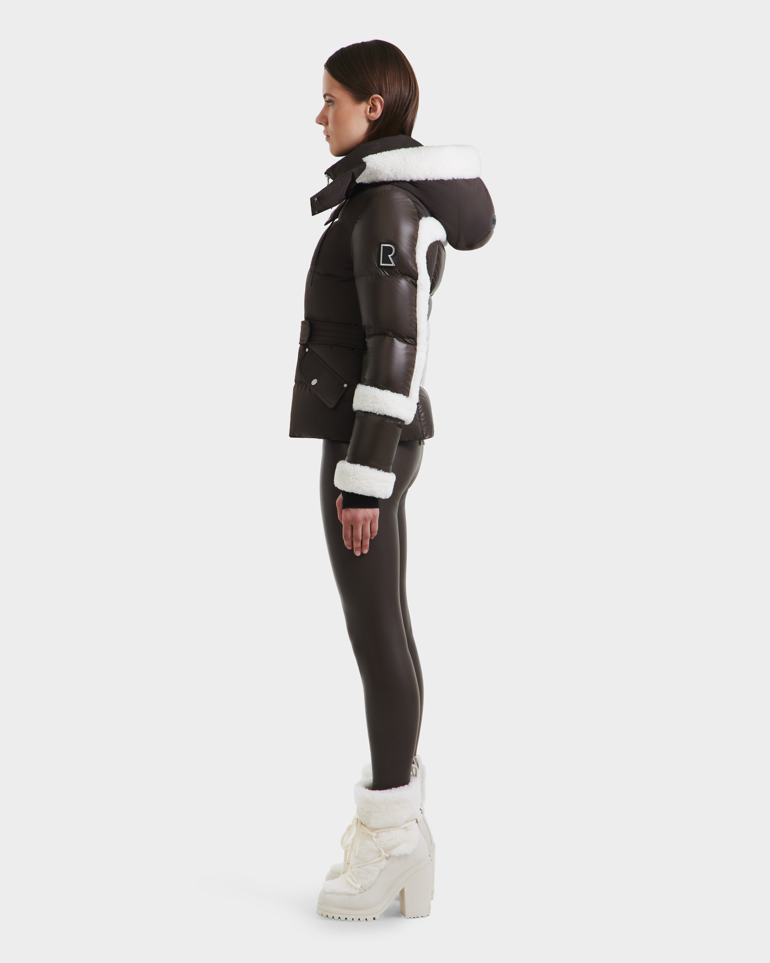 A woman stands in profile wearing the Rudsak AUDREY - 8124582 Espresso belted puffer jacket with white faux fur trim, paired with black leggings and white heeled winter boots with fur.