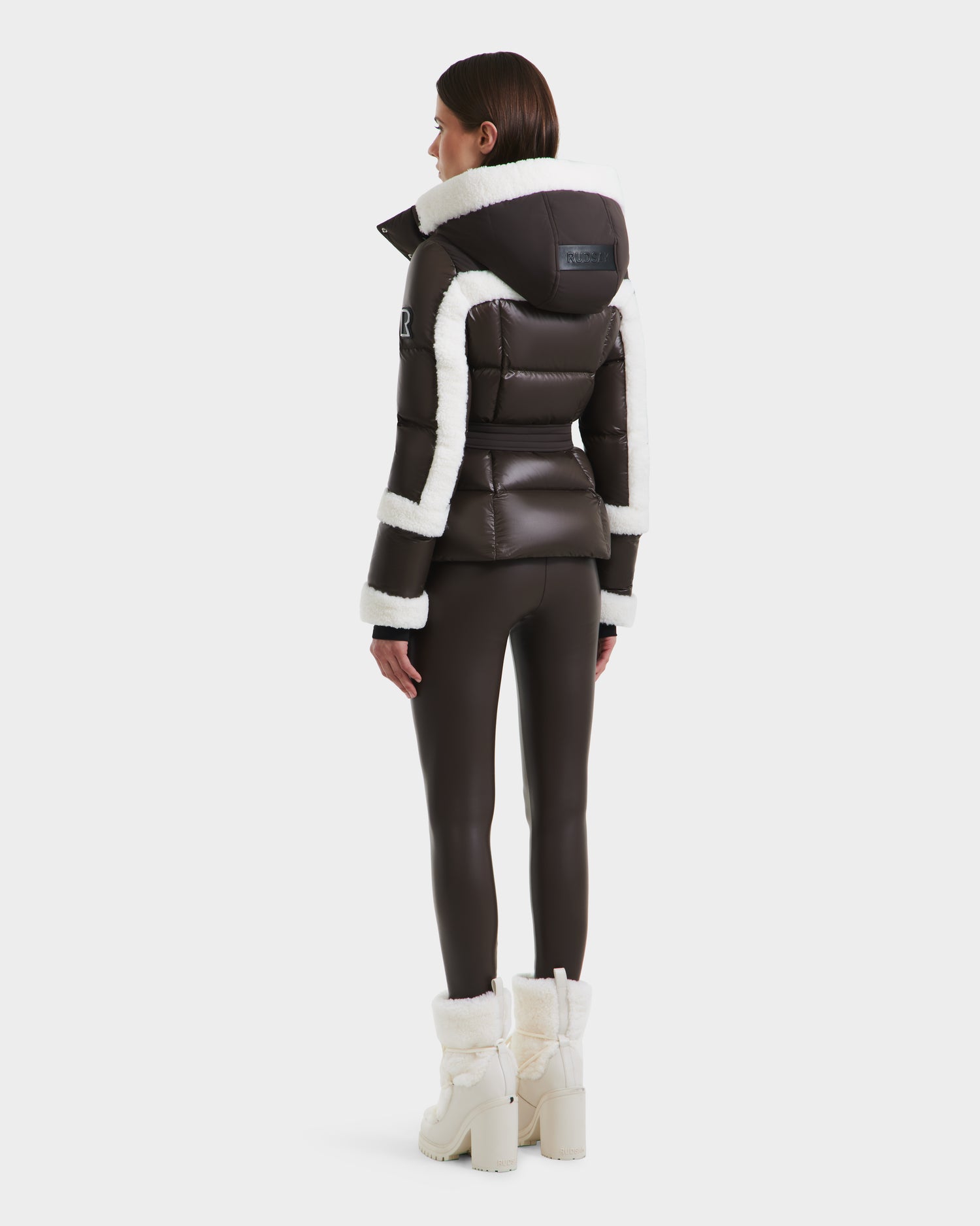 A woman faces away, wearing the Rudsak AUDREY - 8124582 Espresso fitted black down puffer jacket with white faux fur trim, paired with matching black leggings and white fur-lined heeled boots against a plain white background.