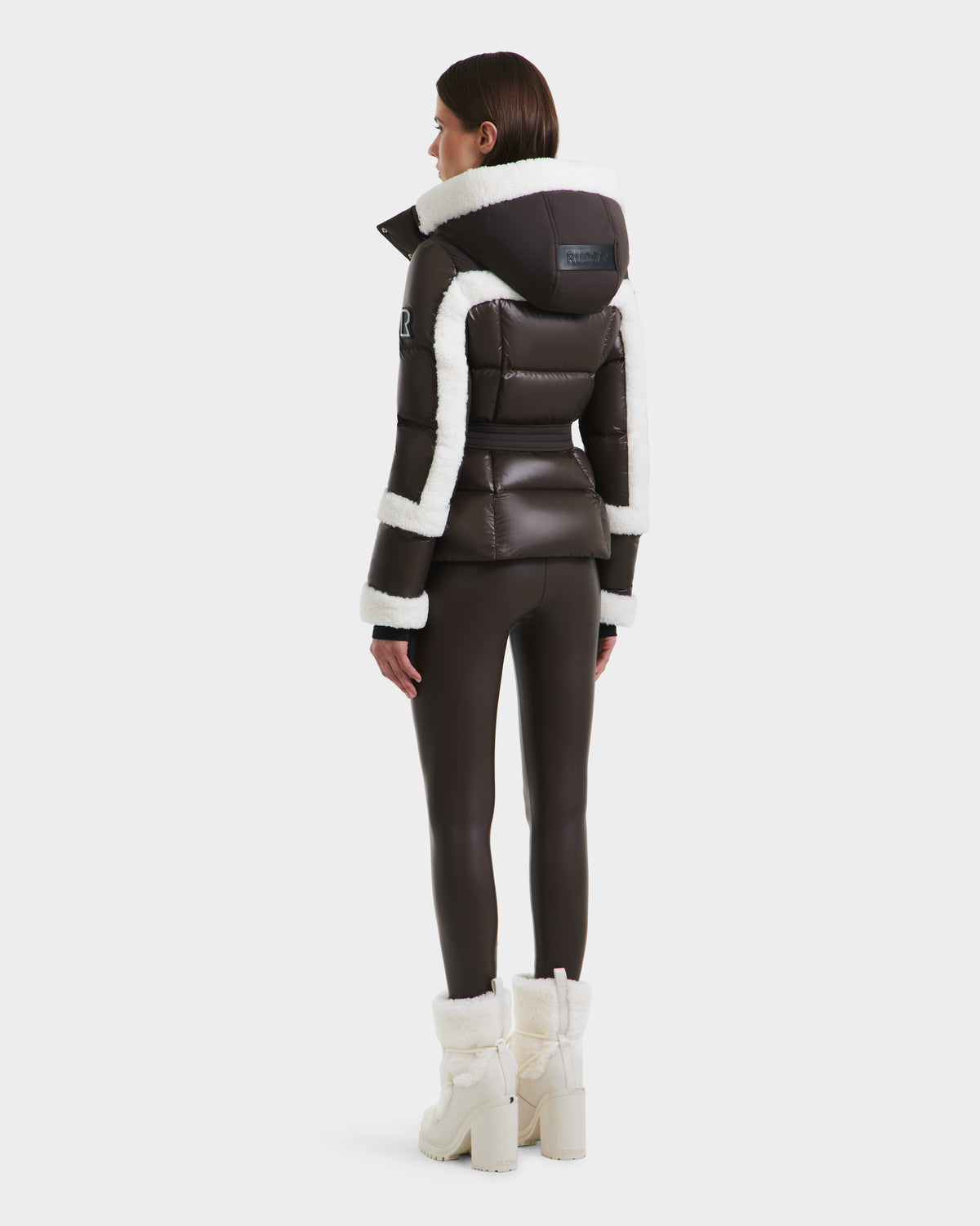 A woman faces away, wearing the Rudsak AUDREY - 8124582 Espresso fitted black down puffer jacket with white faux fur trim, paired with matching black leggings and white fur-lined heeled boots against a plain white background.
