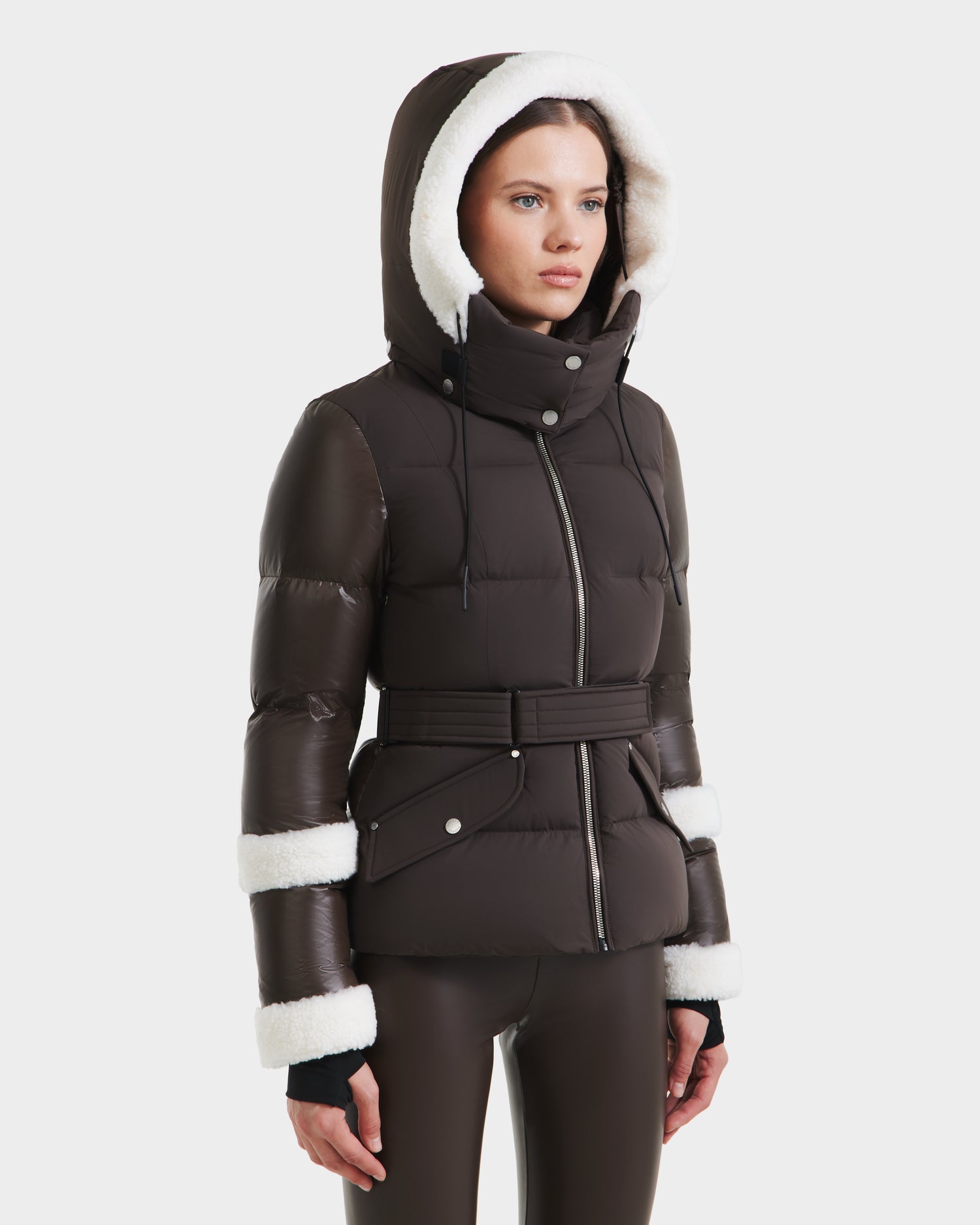 A woman wears the AUDREY - 8124582 Espresso puffer jacket by Rudsak, featuring RDS certified down, a dark brown belted design, white faux fur hood trim, zip and snap closures, styled with matching brown leggings against a plain white background.