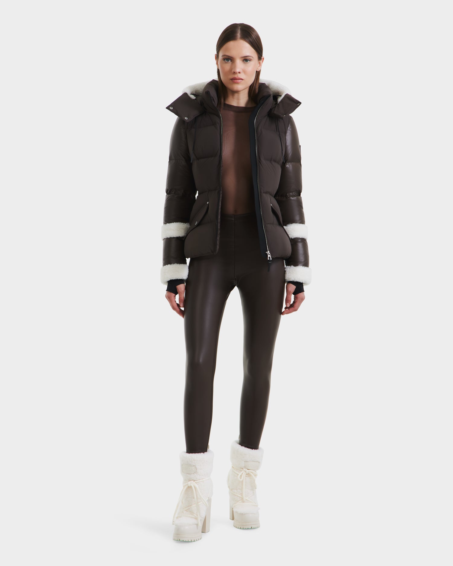 A woman faces forward wearing the Rudsak AUDREY - 8124582 Espresso, a shiny black down puffer with white fur trim, sheer black top, leggings, and white fur-trimmed boots. The jacket uses RDS certified down for ethical warmth. Plain white background.