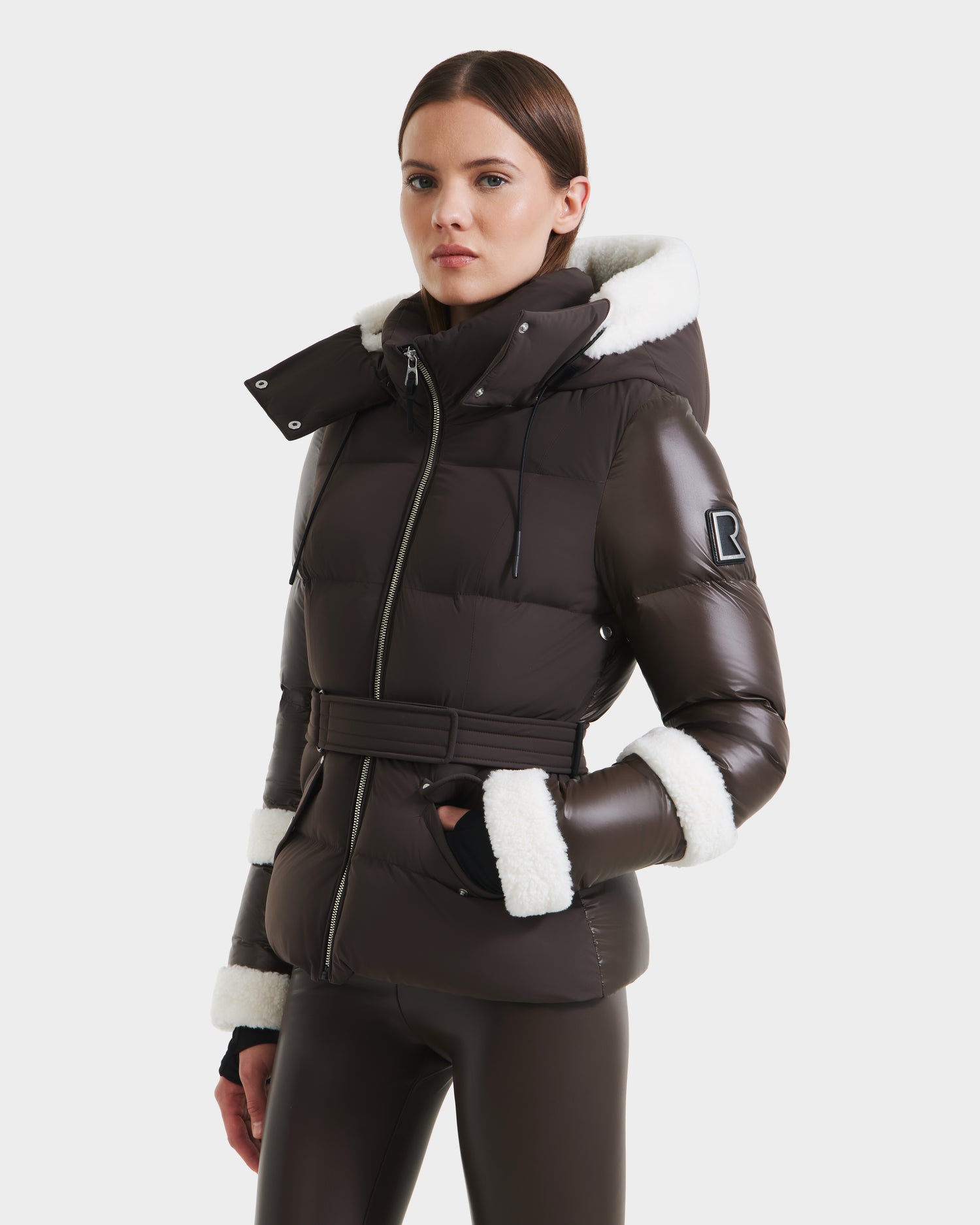 A woman in the Rudsak AUDREY - 8124582 Espresso, a dark brown belted down puffer jacket with white faux fur trim made from recycled Stormshell fabric, is paired with matching pants against a white background.