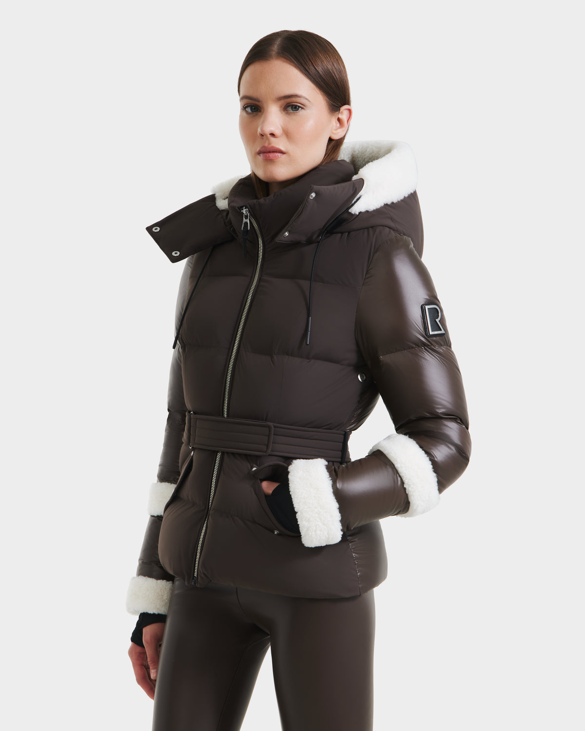 A woman in the Rudsak AUDREY - 8124582 Espresso, a dark brown belted down puffer jacket with white faux fur trim made from recycled Stormshell fabric, is paired with matching pants against a white background.
