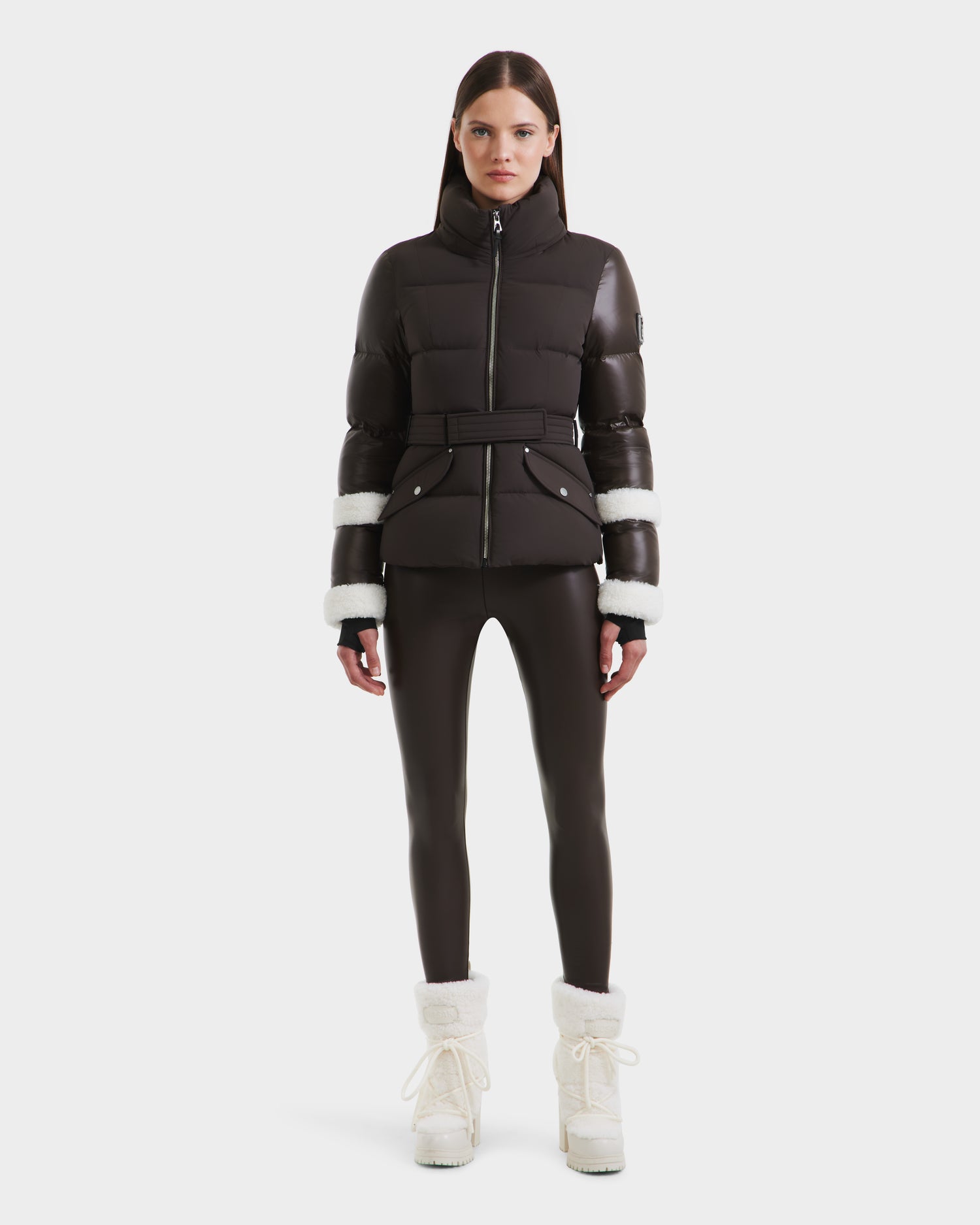 A woman with straight brown hair and a neutral expression faces forward, wearing the Rudsak AUDREY - 8124582 Espresso down puffer jacket, black fitted pants, and white winter boots.