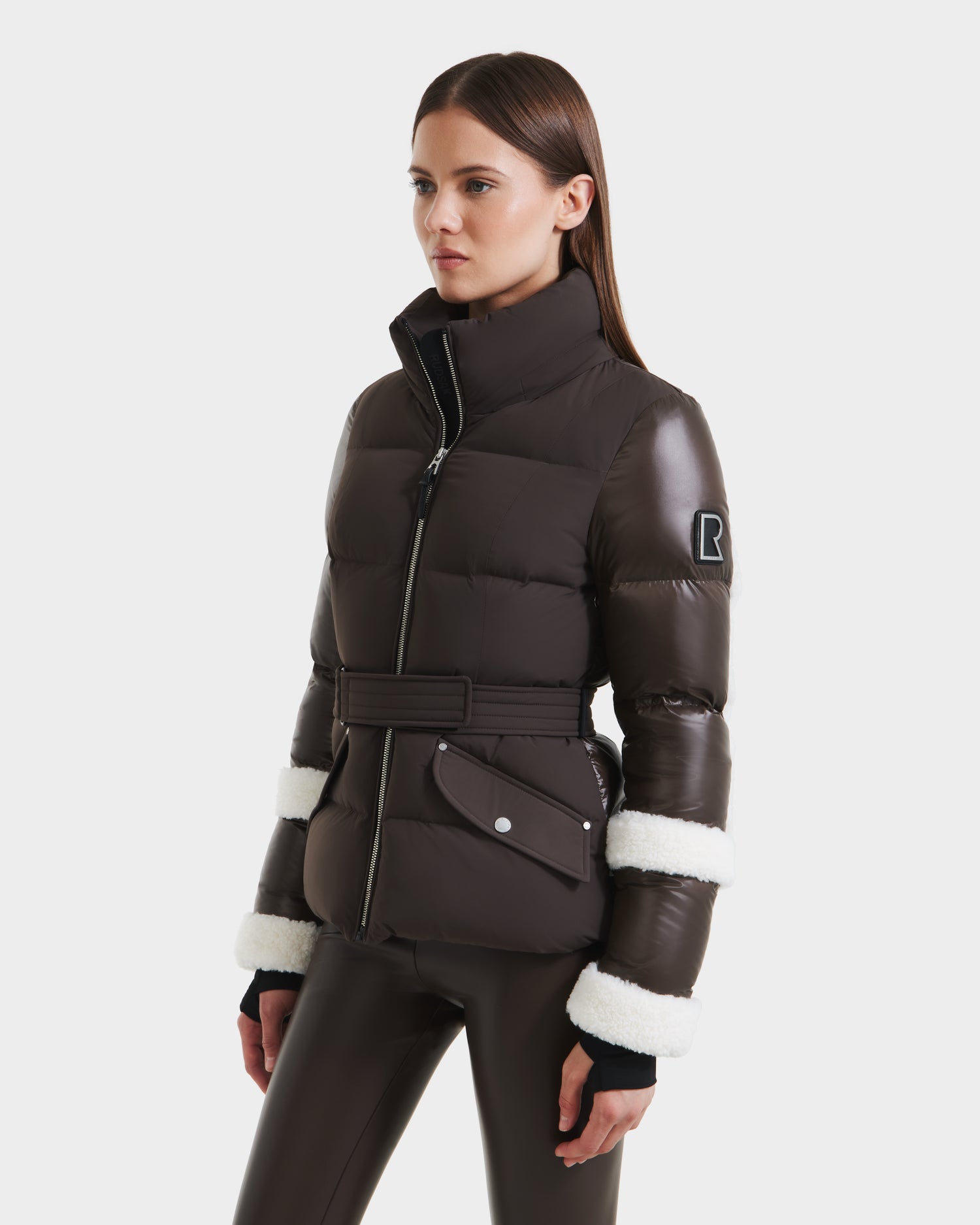 A woman wears the Rudsak AUDREY - 8124582 Espresso dark brown puffer jacket, featuring white faux fur sleeve trim, high collar, belted waist, zipped pockets, and recycled Stormshell fabric—styled with matching pants.