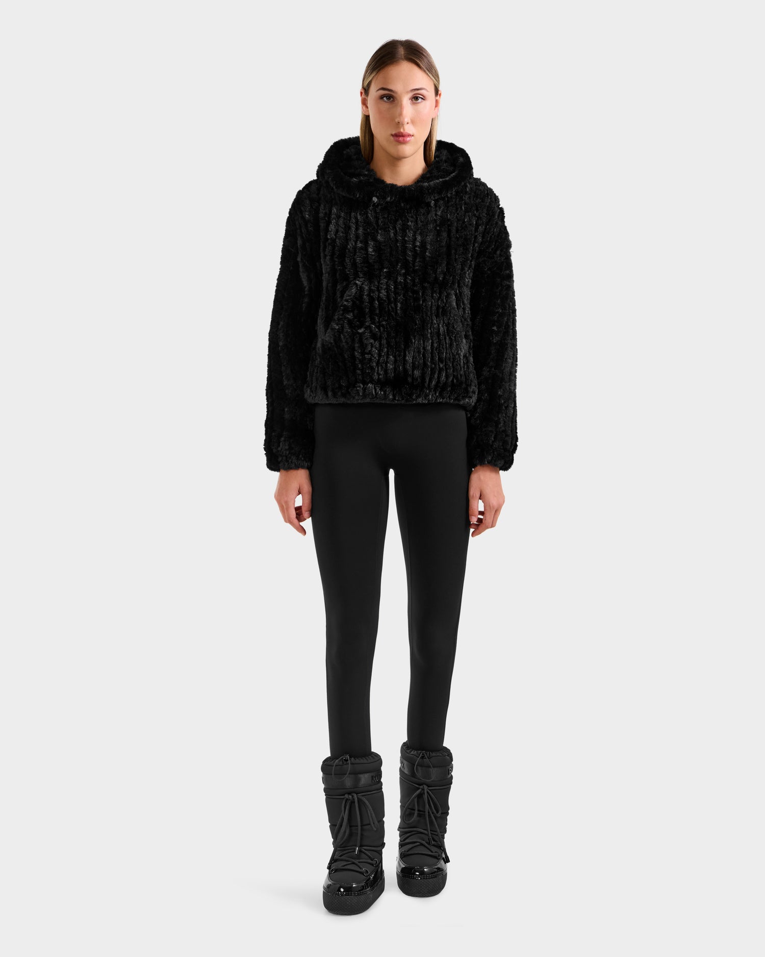 A person faces forward in the Rudsak AUGUST - 8525658 Black faux fur hoodie, paired with black leggings and winter boots—ideal for cozy winter lounging against a plain white background.