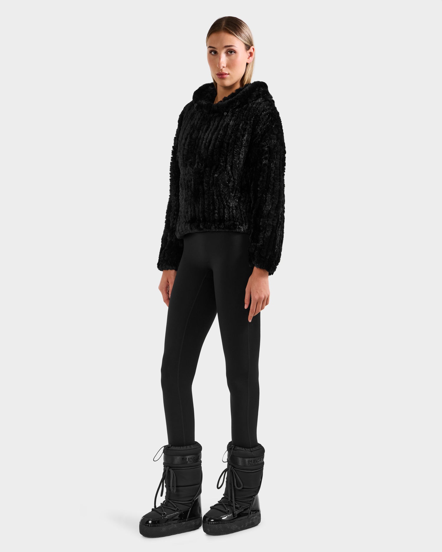 A woman faces forward in a Rudsak AUGUST - 8525658 Black faux fur hoodie, ideal for winter lounging, paired with black leggings and moon boots against a plain light gray background.