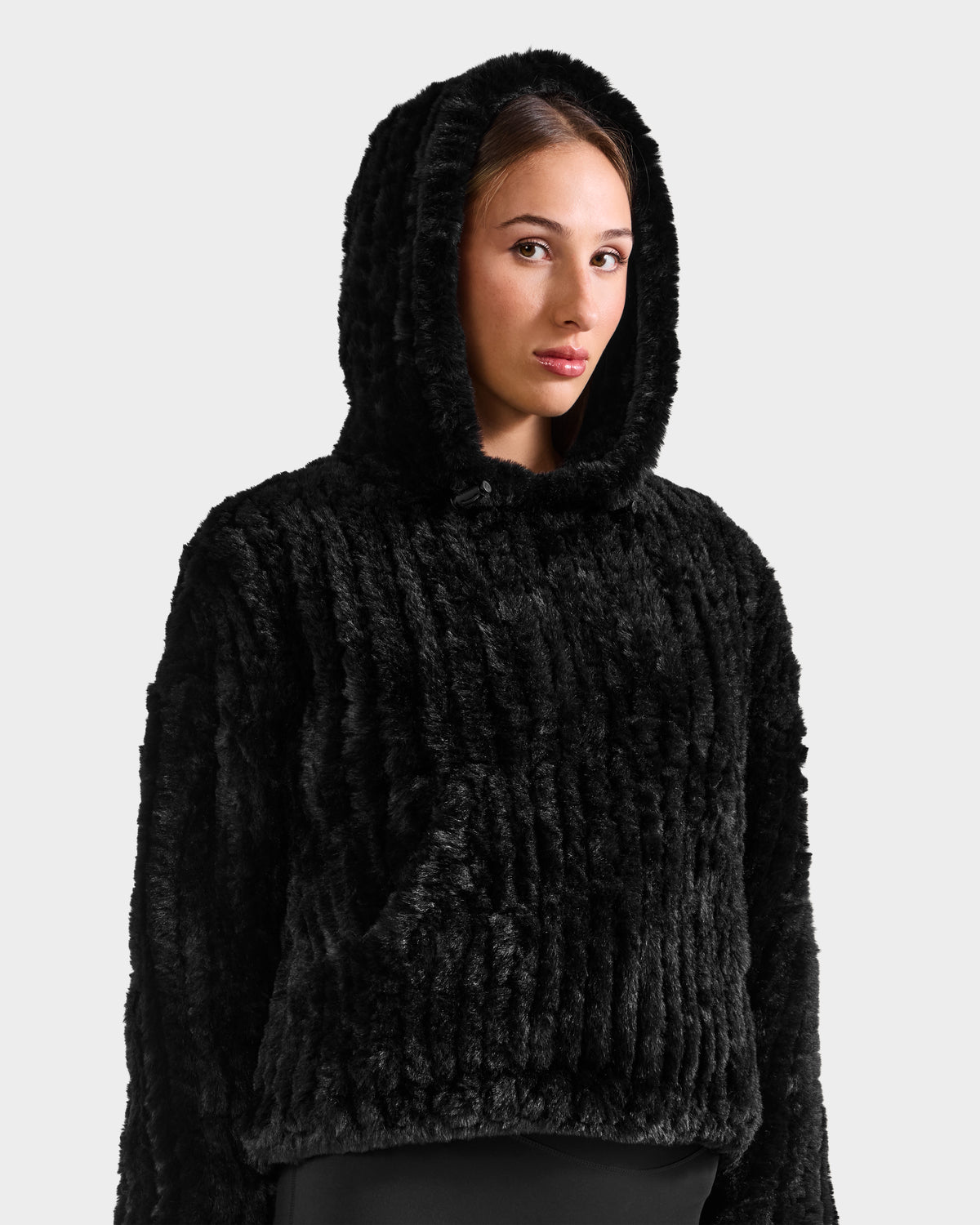A woman models the Rudsak AUGUST - 8525658 Black textured faux fur hoodie, exuding cozy winter lounge style against a plain light gray background.