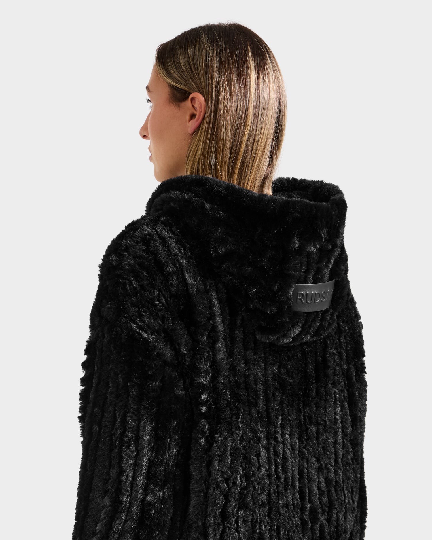 A person with straight, light brown hair is seen from behind in a textured black Rudsak AUGUST - 8525658 faux fur hoodie with a raised collar and branded leather patch, ideal for après-ski or winter lounging. The background is plain and light-colored.
