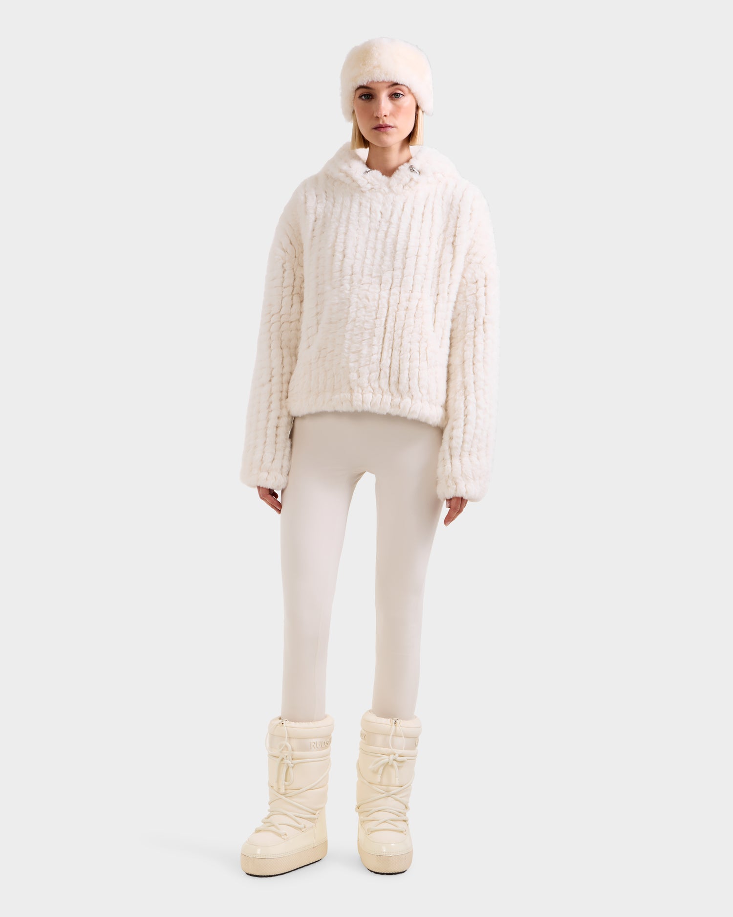 A woman faces forward in Rudsak’s AUGUST - 8525658 Iceland: an off-white faux fur hat, matching jacket, cream leggings, and boots—perfect for après-ski style against a plain light background.