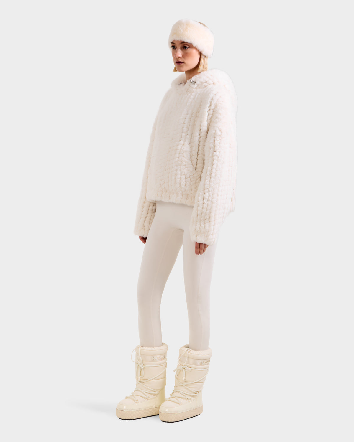 A model wears the AUGUST - 8525658 Iceland faux fur knit hoodie by Rudsak with a furry white hood, headband, cream leggings, and beige winter boots—ideal for après-ski or winter lounging against a plain white background.