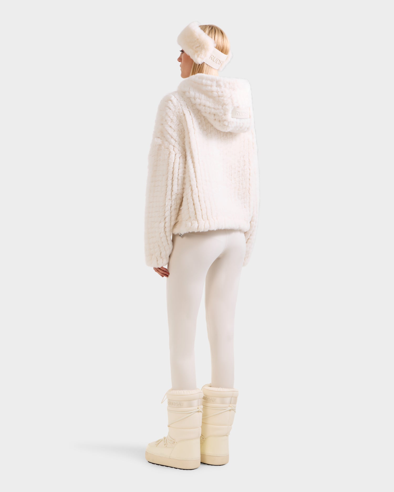 A person stands facing away, wearing Rudsak’s AUGUST - 8525658 Iceland white faux fur knit hoodie, paired with white leggings, cream snow boots, and a matching headband—ideal for après-ski or cozy winter lounging.