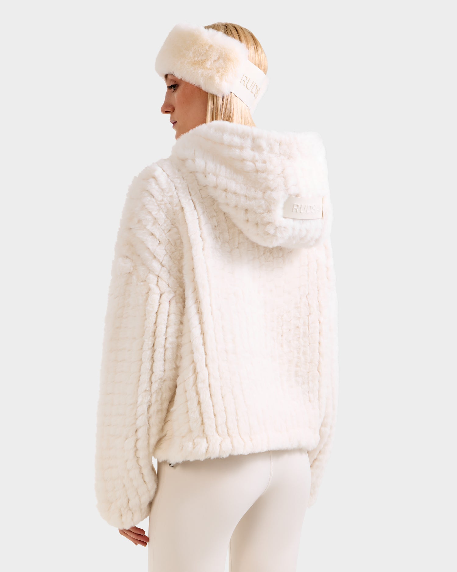 A person faces away in a Rudsak AUGUST - 8525658 Iceland fluffy faux fur knit hoodie with matching white headband, highlighting the textured material. Paired with white leggings, the winter-ready set stands out against a plain light background.