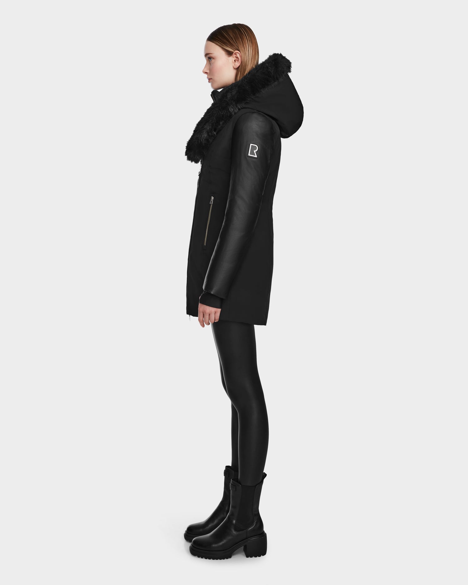 A woman stands in profile against a white background wearing the Rudsak AURELLE - L124538 Black women’s down parka with vegan leather sleeves, faux fur trim, and zippered pockets, styled with black leggings and boots.