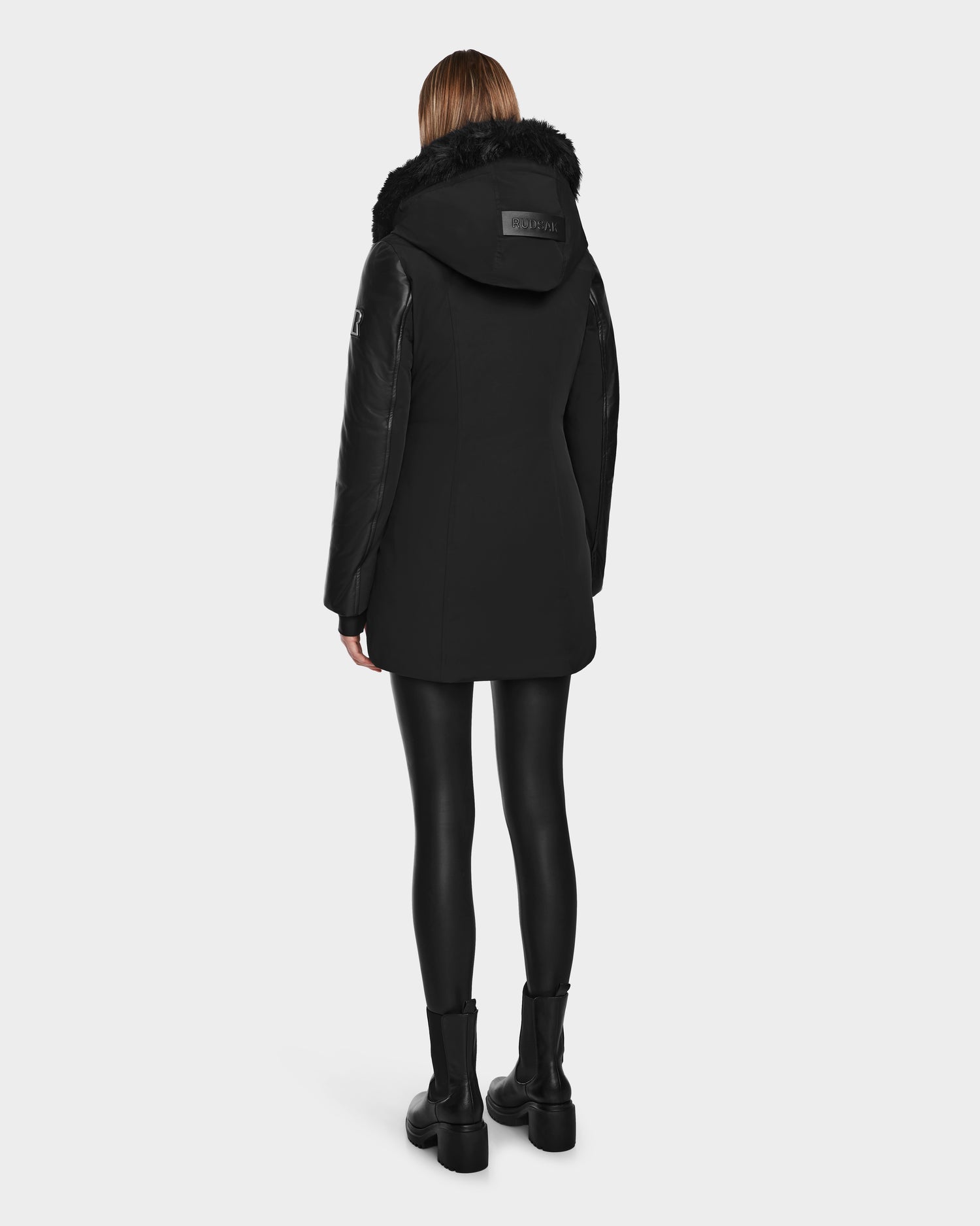 A woman with straight, light brown hair faces away, wearing Rudsak’s AURELLE - L124538 Black down parka with faux fur trim and vegan leather sleeves, paired with black leggings and boots against a plain white background.