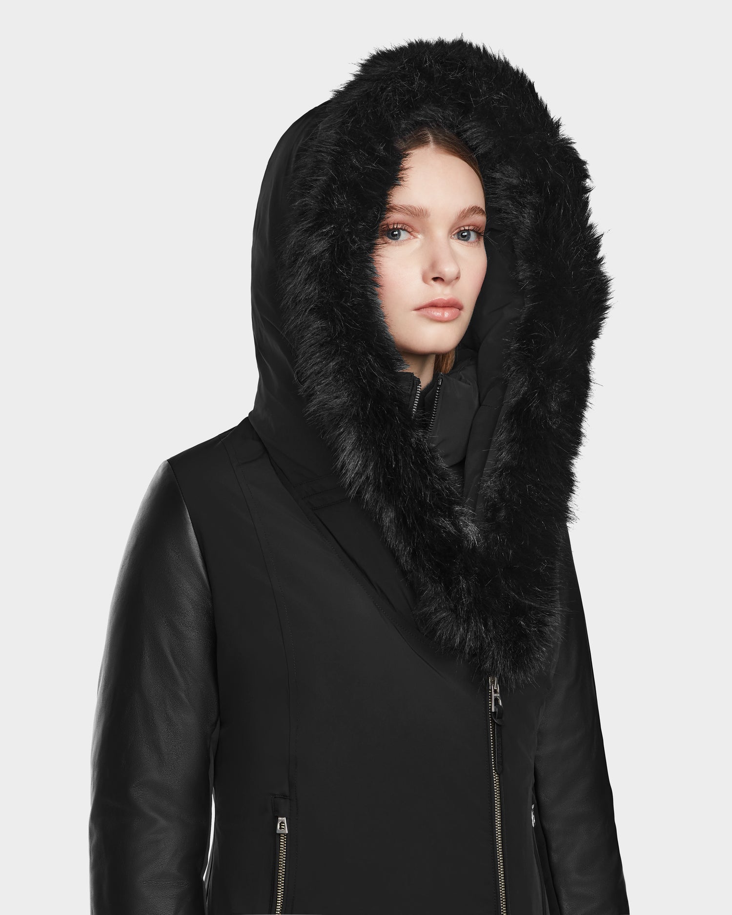 A young woman models the Rudsak AURELLE - L124538 Black parka, featuring vegan leather sleeves and a large faux fur-trimmed hood, standing against a plain light background and looking slightly to the side.