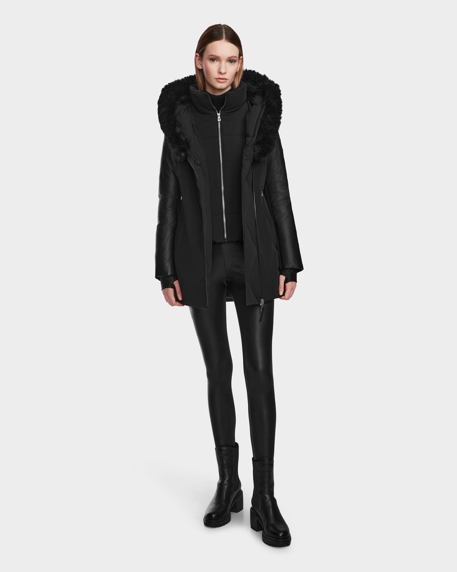 A person stands facing forward wearing the Rudsak AURELLE - L124538 Black hooded coat with faux fur trim and vegan leather sleeves, black zip-up top, leggings, and heeled boots against a plain white background.