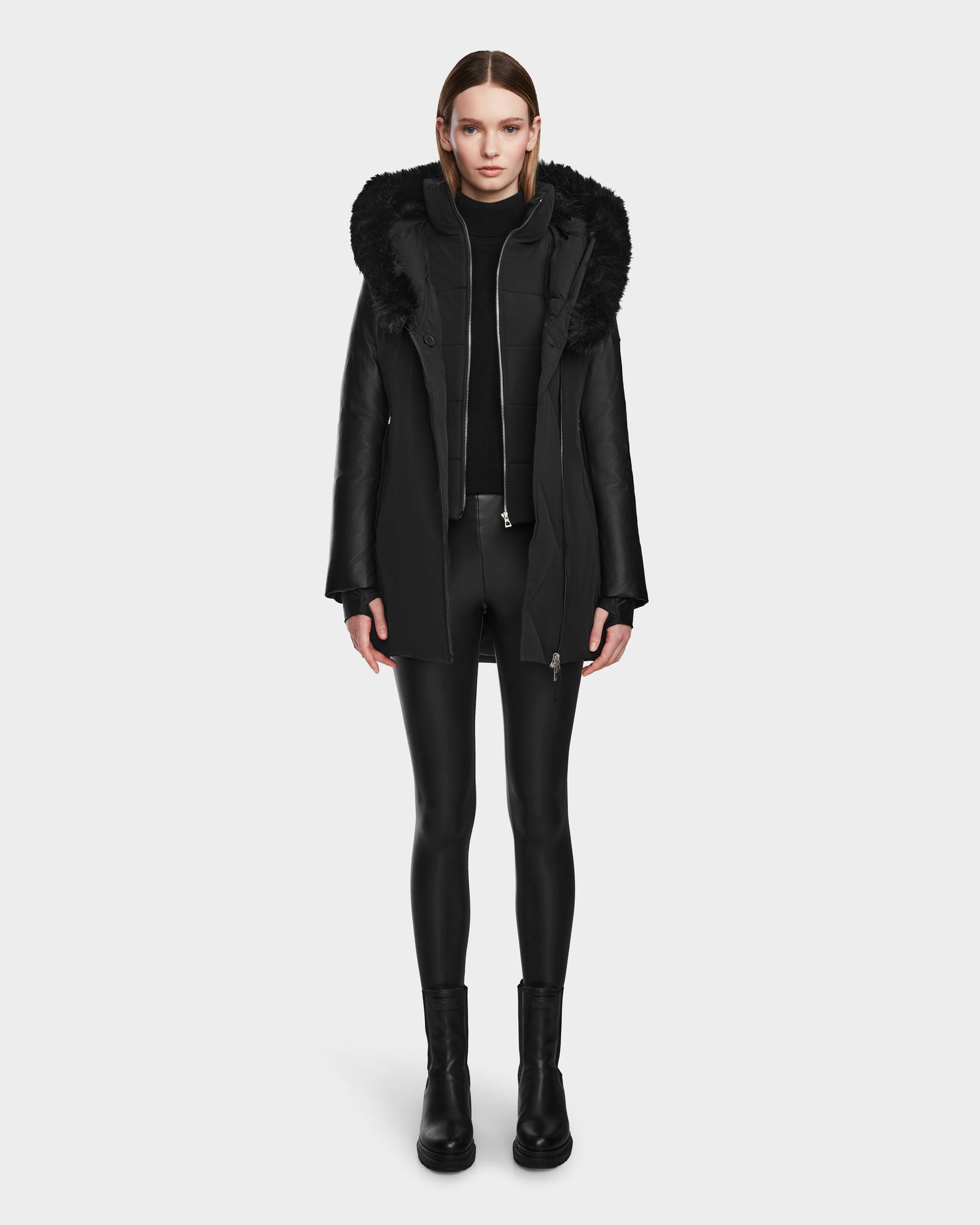 A woman faces forward wearing the Rudsak AURELLE - L124538 Black women’s down parka with faux fur trim, paired with a black turtleneck, leggings, and ankle boots against a plain white background.