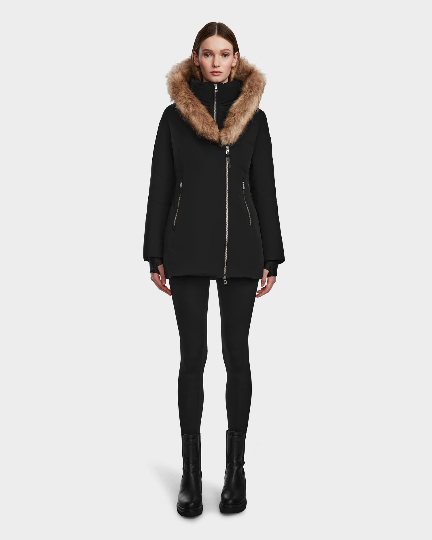 A woman faces forward against a white background wearing the Rudsak AURORE - L123561 Black/Natural fitted down parka with a large brown faux fur hood, paired with black leggings and black boots.