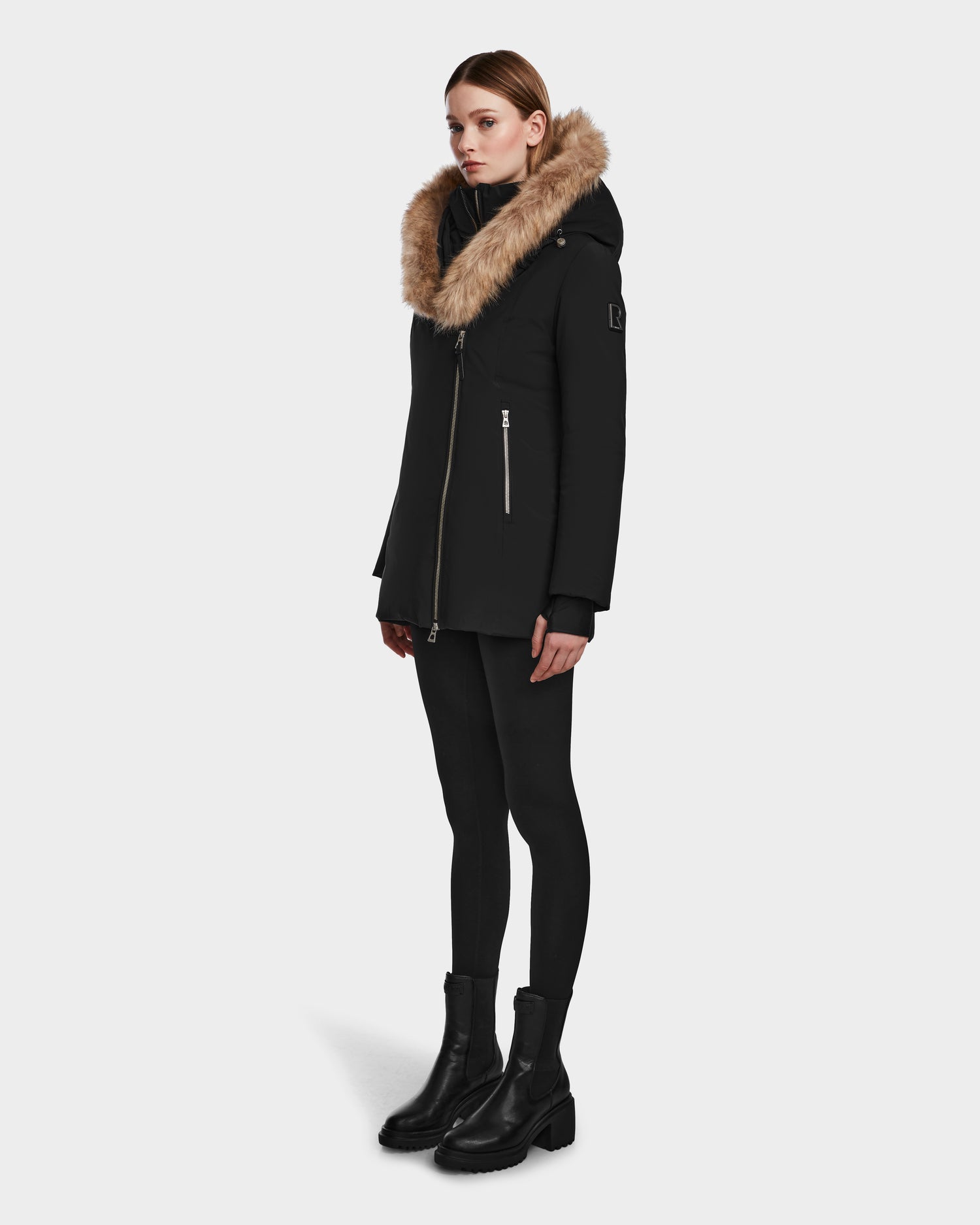 A woman wears the Rudsak AURORE - L123561 Black/Natural fitted down parka with a large tan faux fur hood, paired with black leggings and ankle boots, standing against a plain light gray background.