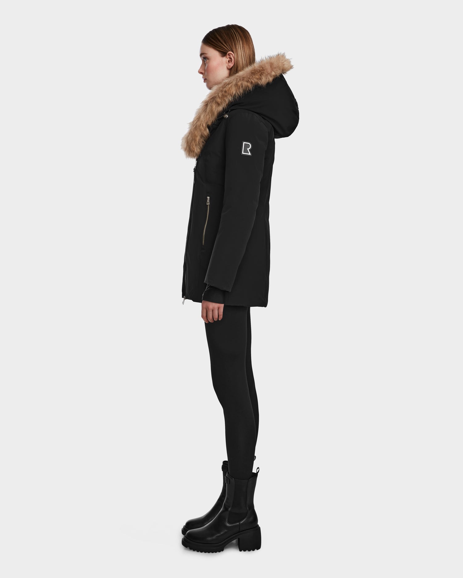 A woman in the Rudsak AURORE - L123561 Black/Natural down parka with a fur-trimmed hood, black leggings, and boots stands in profile against a white background. The weather-resistant parka has a zip closure and a logo patch on the upper sleeve.
