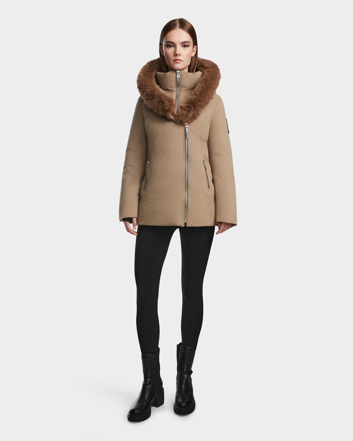 A woman faces forward against a white background, wearing the Rudsak AURORE - L123561 Sand fitted down parka with a large brown fur collar, along with black leggings and black boots.