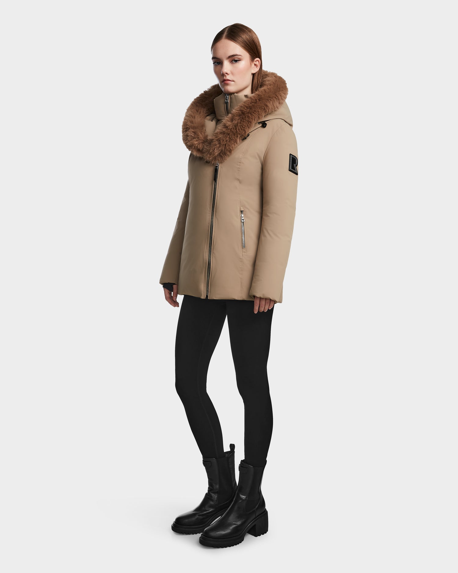 A woman stands wearing the Rudsak AURORE - L123561 Sand women's down parka with a large fur collar, paired with black leggings and chunky black boots, against a plain white background.
