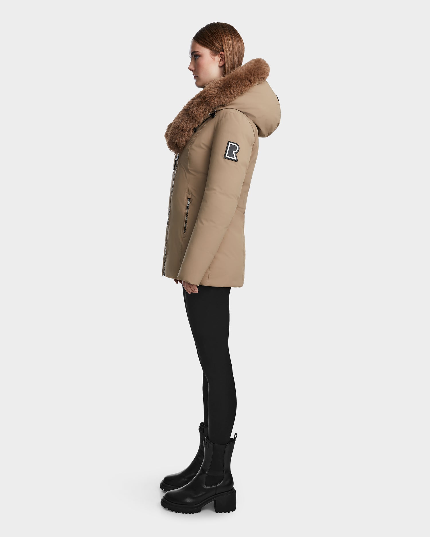 A woman stands in profile wearing the Rudsak AURORE - L123561 Sand fitted down parka with a large brown fur-trimmed hood, black leggings, and boots. The jacket has a zipper and upper arm patch. A white background highlights her look.