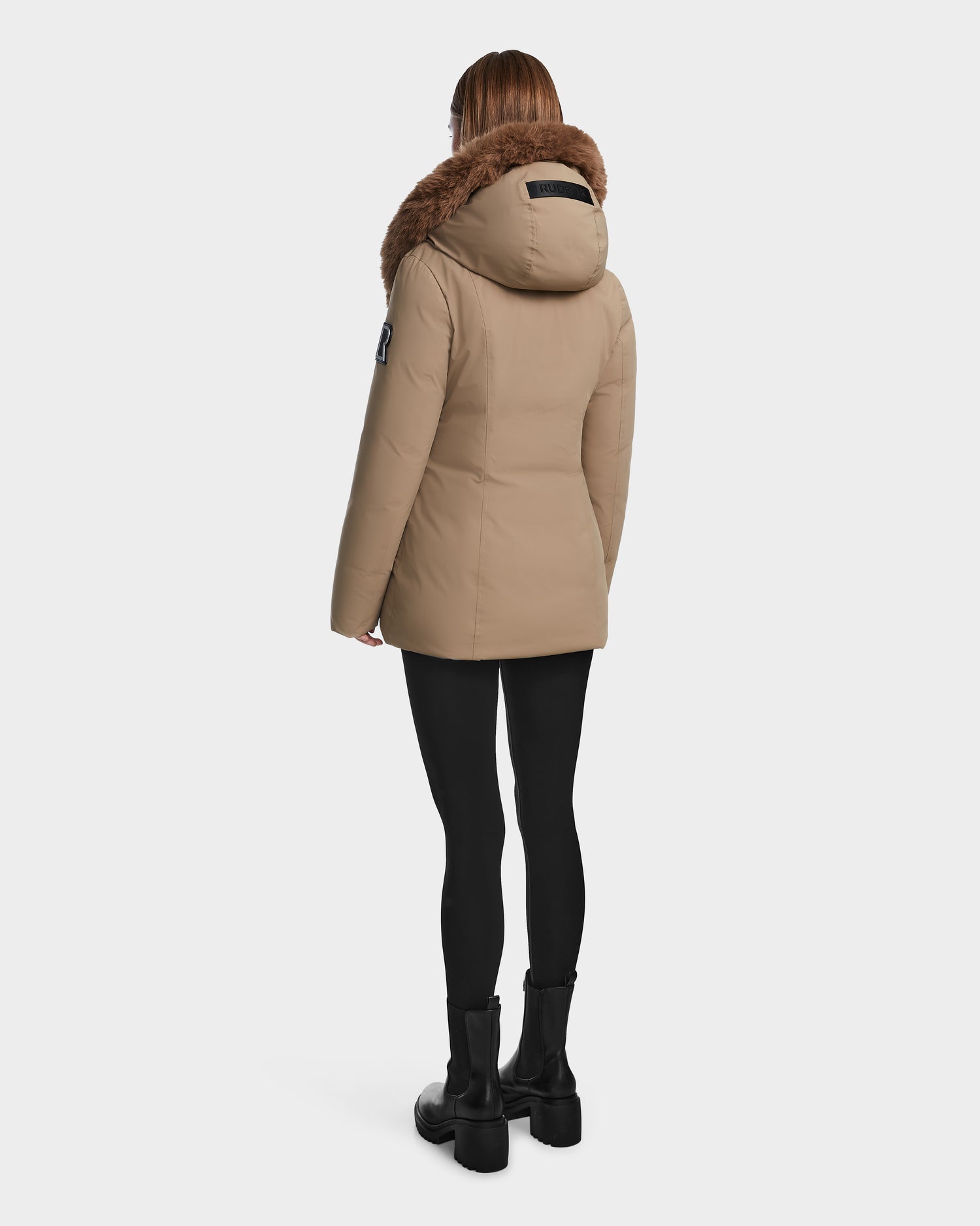 A person stands with their back to the camera, wearing the Rudsak AURORE - L123561 Sand fitted down parka with brown fur trim, black leggings, and black ankle boots against a plain white background.