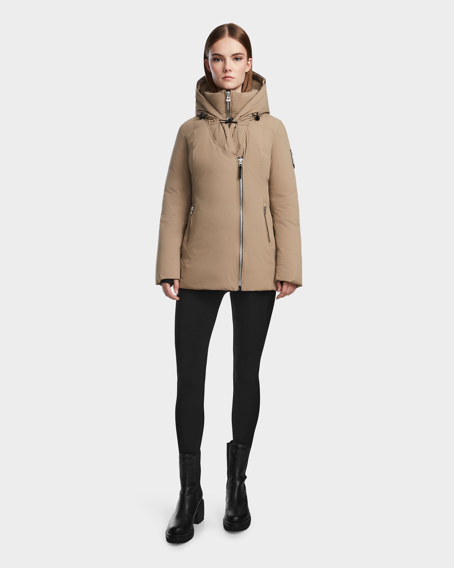 A woman faces forward against a plain white background, wearing the Rudsak AURORE - L123561 Sand fitted down parka, paired with black leggings and black ankle boots.