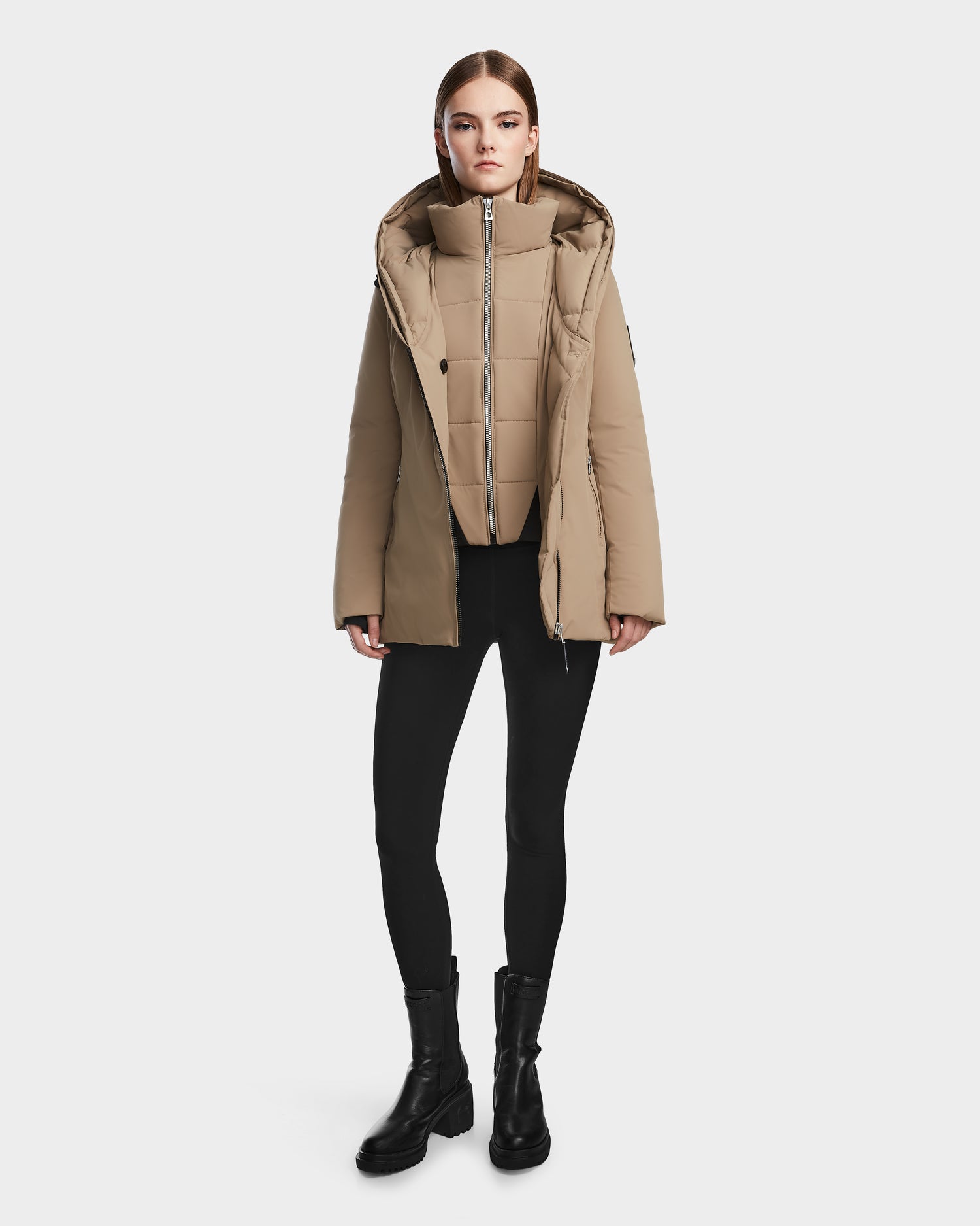 A woman faces forward wearing the Rudsak AURORE - L123561 Sand women's down parka with a hood, paired with black leggings and black ankle boots against a plain light background.
