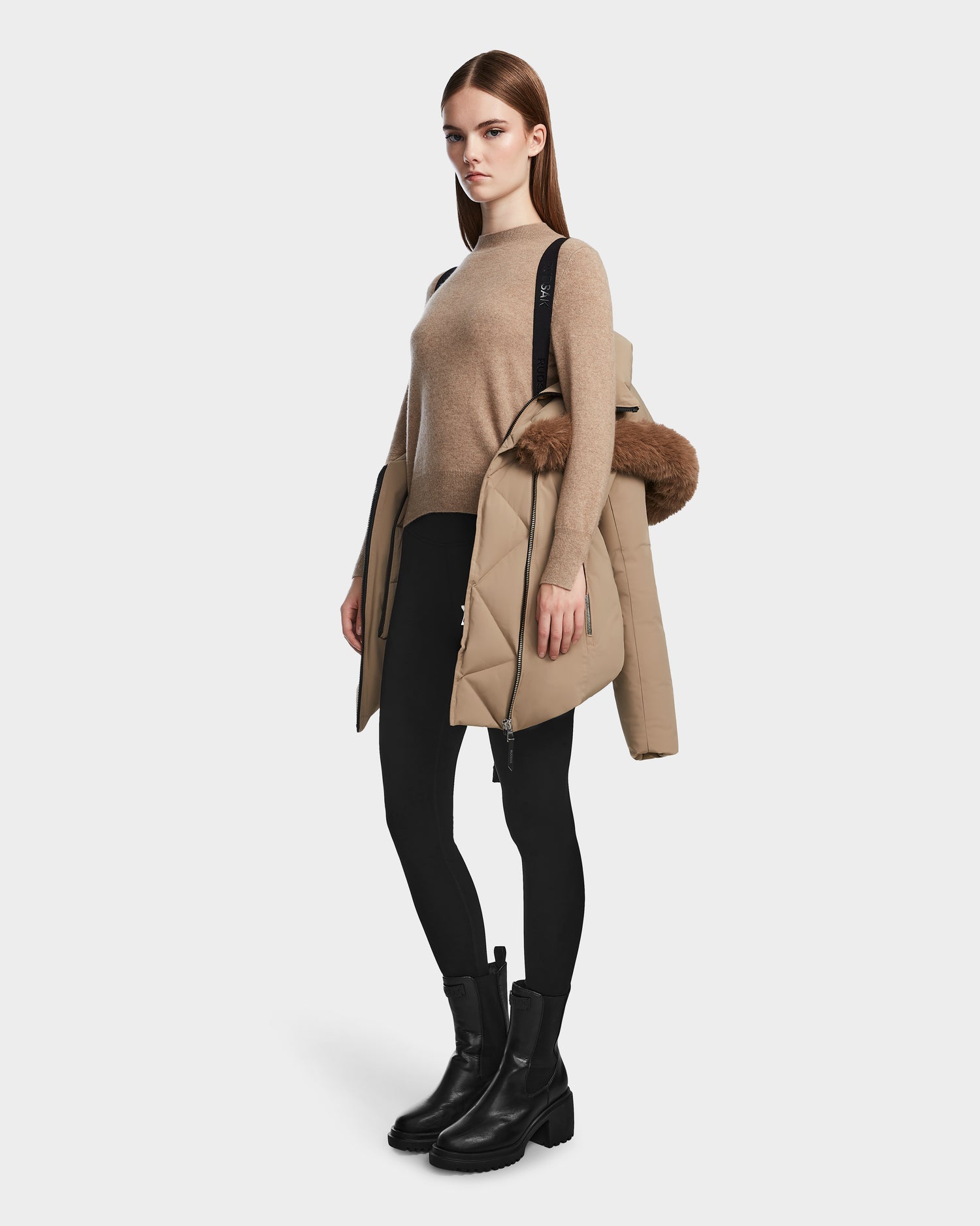 A woman stands against a plain white background holding the Rudsak AURORE - L123561 Sand women's down parka with a fur-trimmed hood draped over her arm, wearing a beige sweater, black leggings, and black boots.