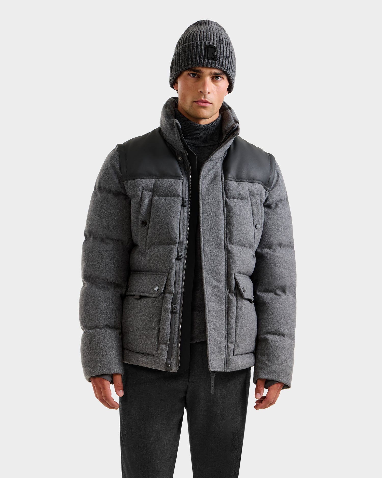 A man stands facing forward against a plain light background, wearing the Rudsak BALDWIN W - 6125532 Heather Charcoal wool-blend parka with black shoulder accents and a grey knit beanie, paired with a dark turtleneck sweater and dark pants.
