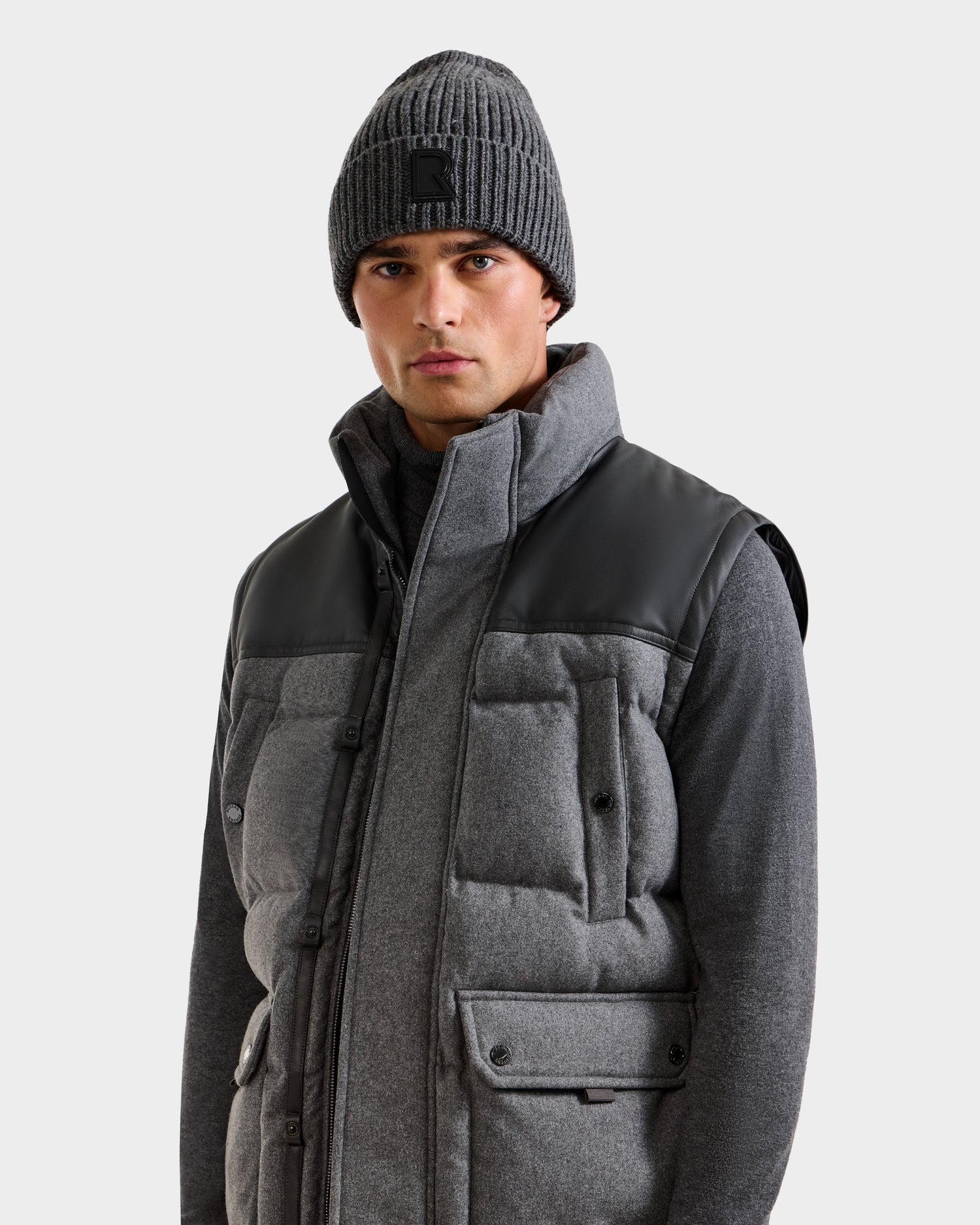A man wearing the Rudsak BALDWIN W - 6125532 Heather Charcoal gray puffer vest with black shoulder panels and a gray zip-up sweater stands against a plain light background.