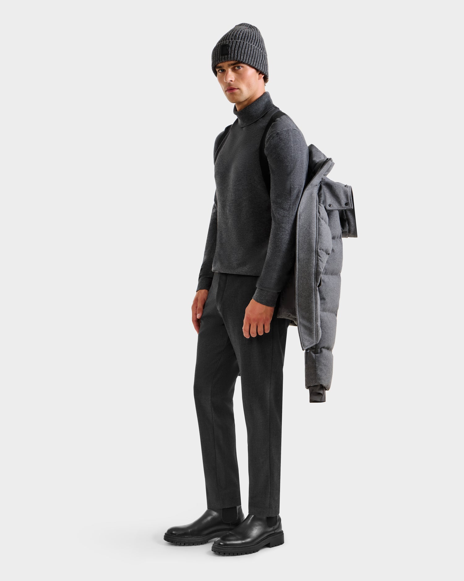 A man stands facing left against a plain white background, wearing dark trousers, a grey sweater, black boots, and a dark knit beanie. He carries the Rudsak BALDWIN W - 6125532 Heather Charcoal men’s down parka draped over his left shoulder.