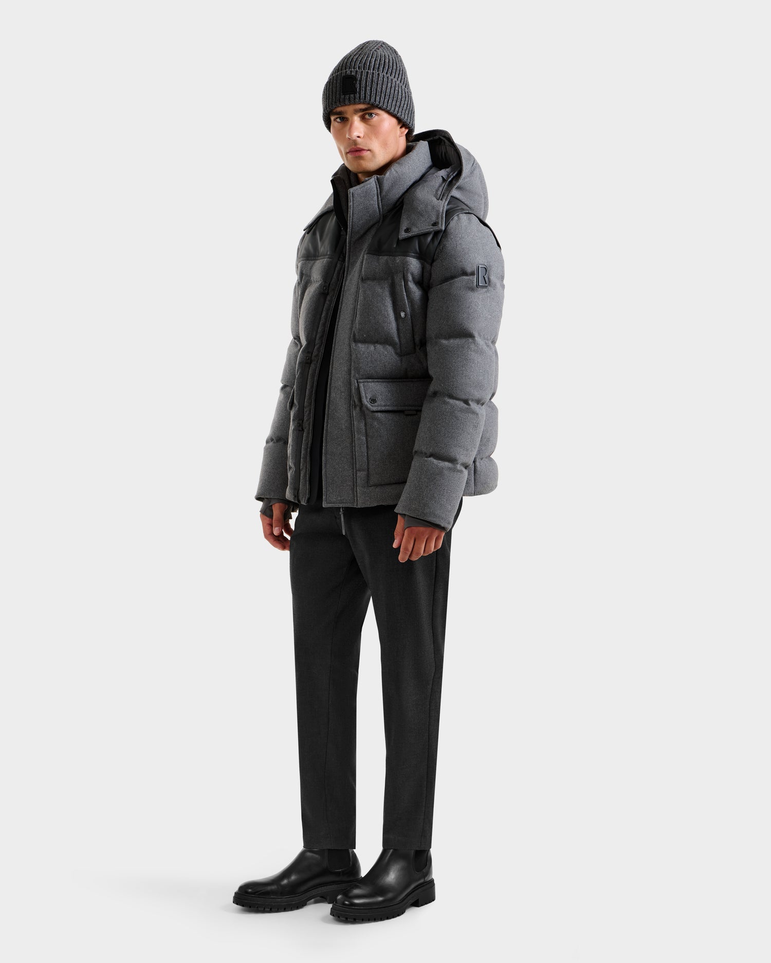 A man showcases the Rudsak BALDWIN W - 6125532 Heather Charcoal men’s down parka with a hood, paired with dark pants, black boots, and a gray knit beanie against a plain white background.