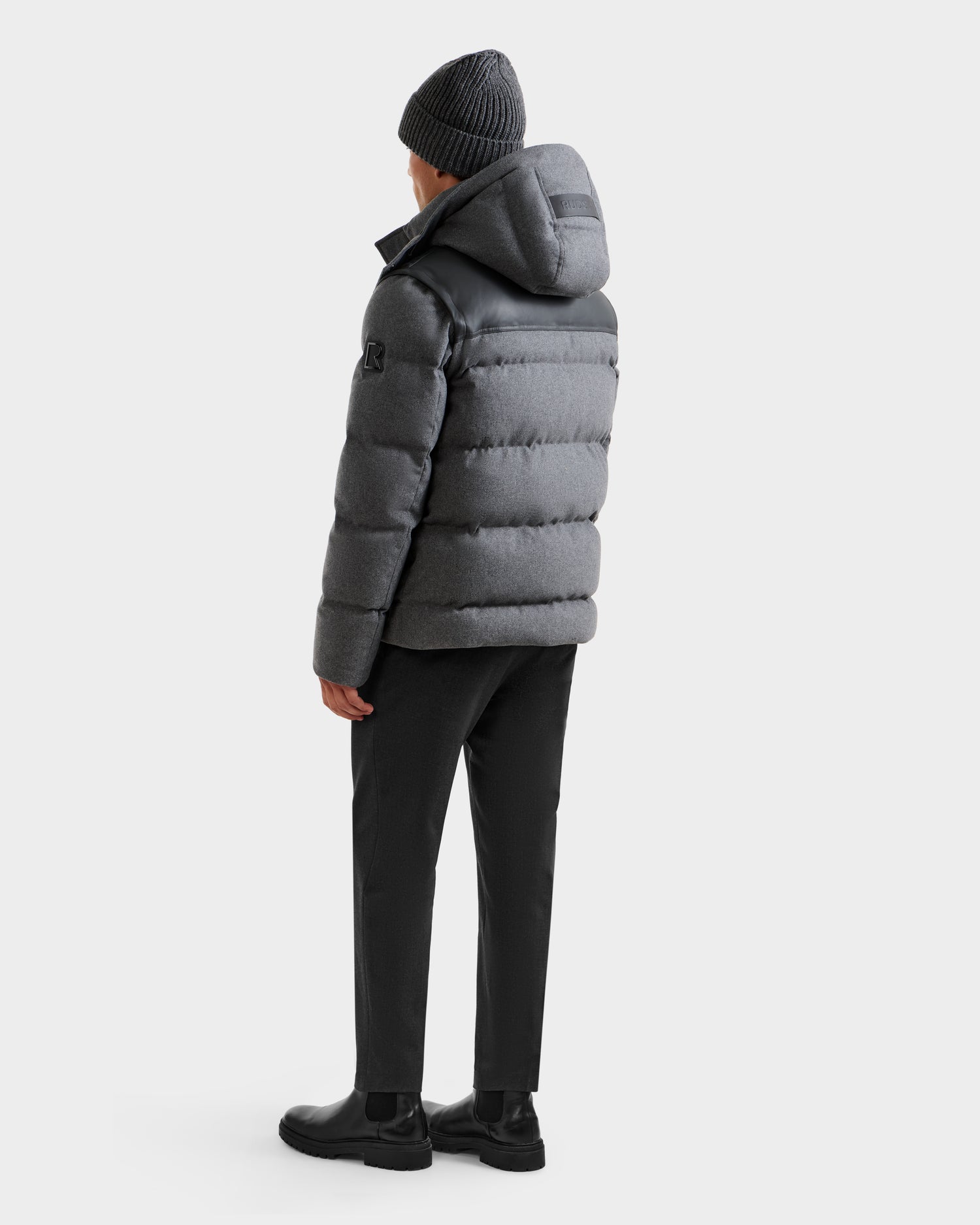 A person stands facing away, wearing the Rudsak BALDWIN W - 6125532 Heather Charcoal men’s down parka with a hood, black beanie, pants, and boots—a perfect winter staple against a plain light background.