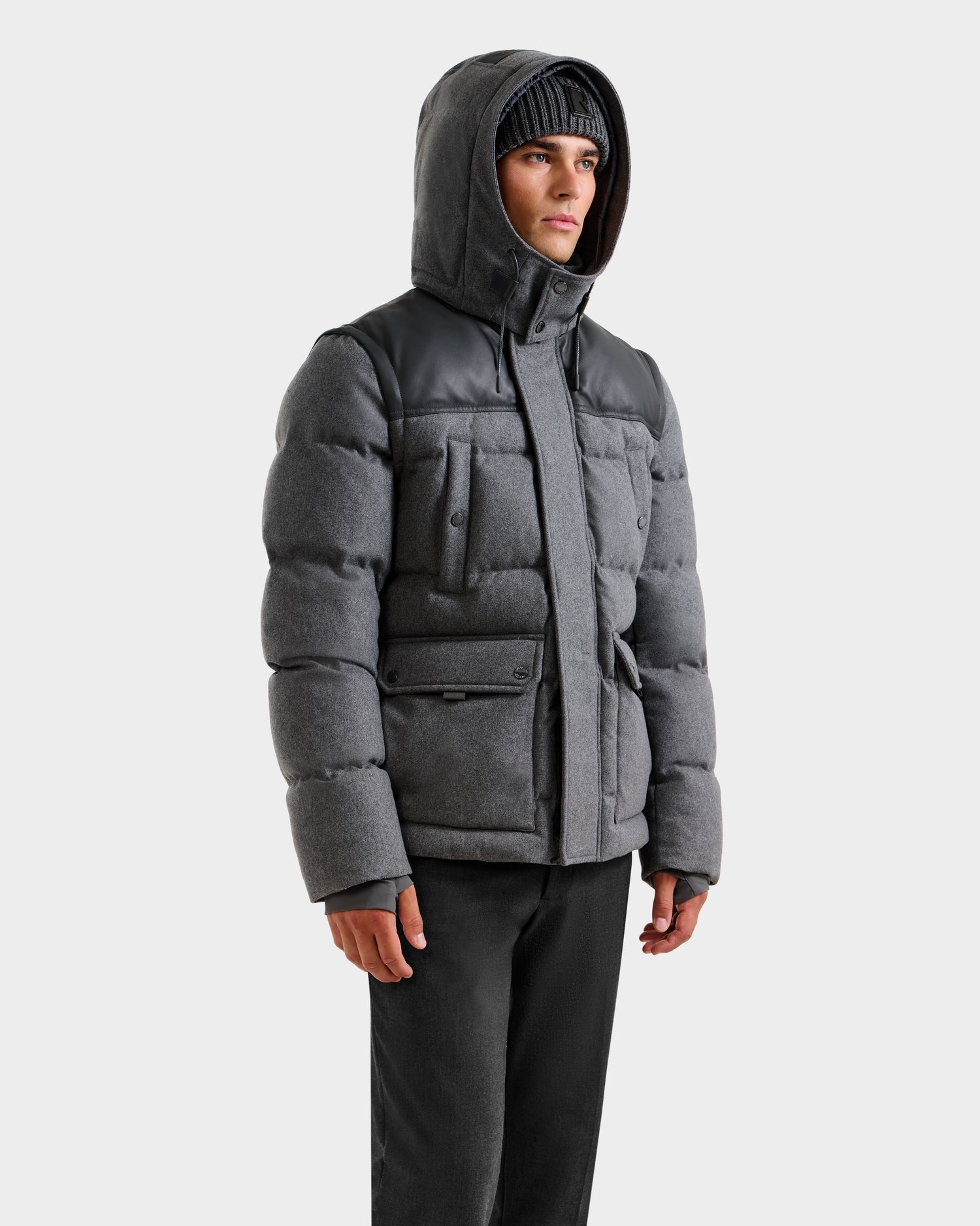 Dressed in the Rudsak BALDWIN W - 6125532 Heather Charcoal men’s down parka with black accents, paired with black pants and a knit beanie, a model stands against a plain white background—an essential winter look.
