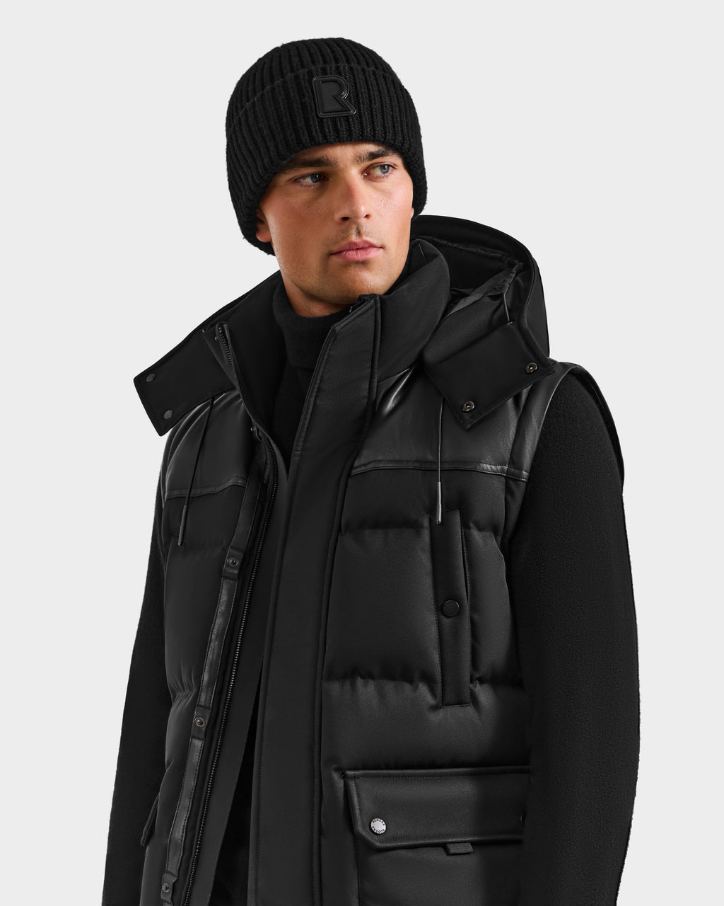 A man wears the Rudsak BALDWIN - 6124507 Black men’s down parka with removable sleeves and snap buttons, styled with a black knit beanie and sweater, gazing to the side against a plain light background.