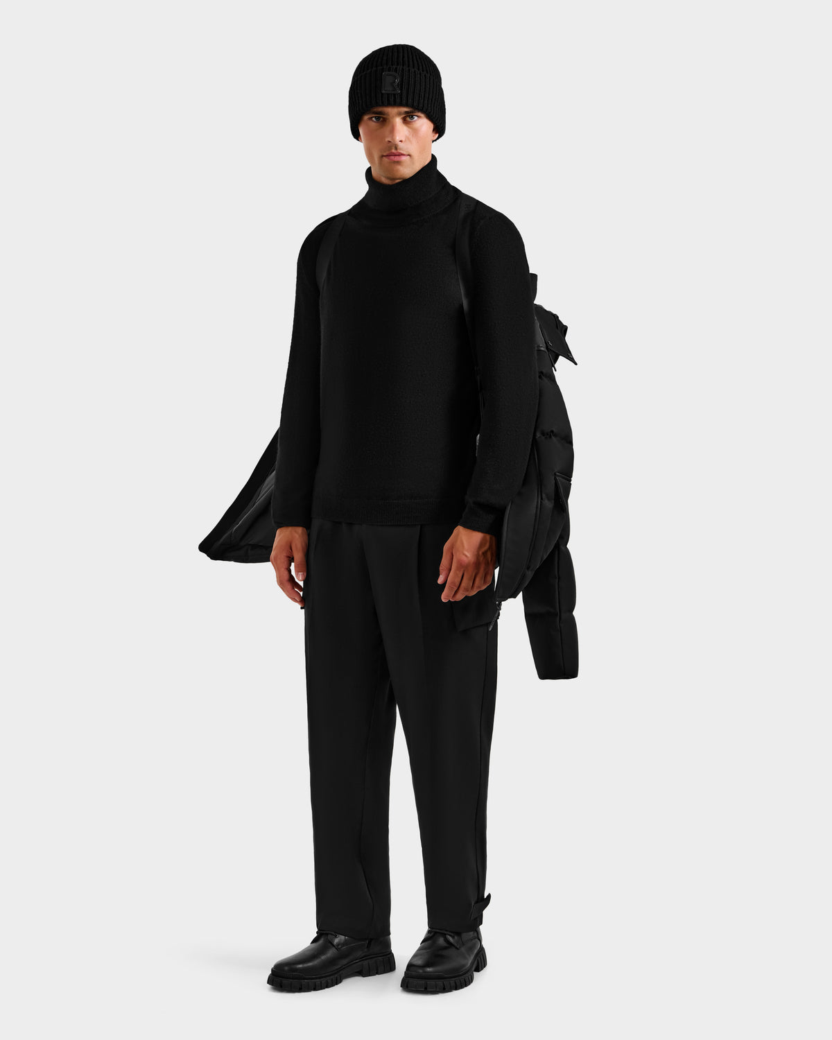 A person in all-black attire holds the Rudsak BALDWIN - 6124507 Black men's down parka with removable sleeves over one shoulder, standing against a plain light background.