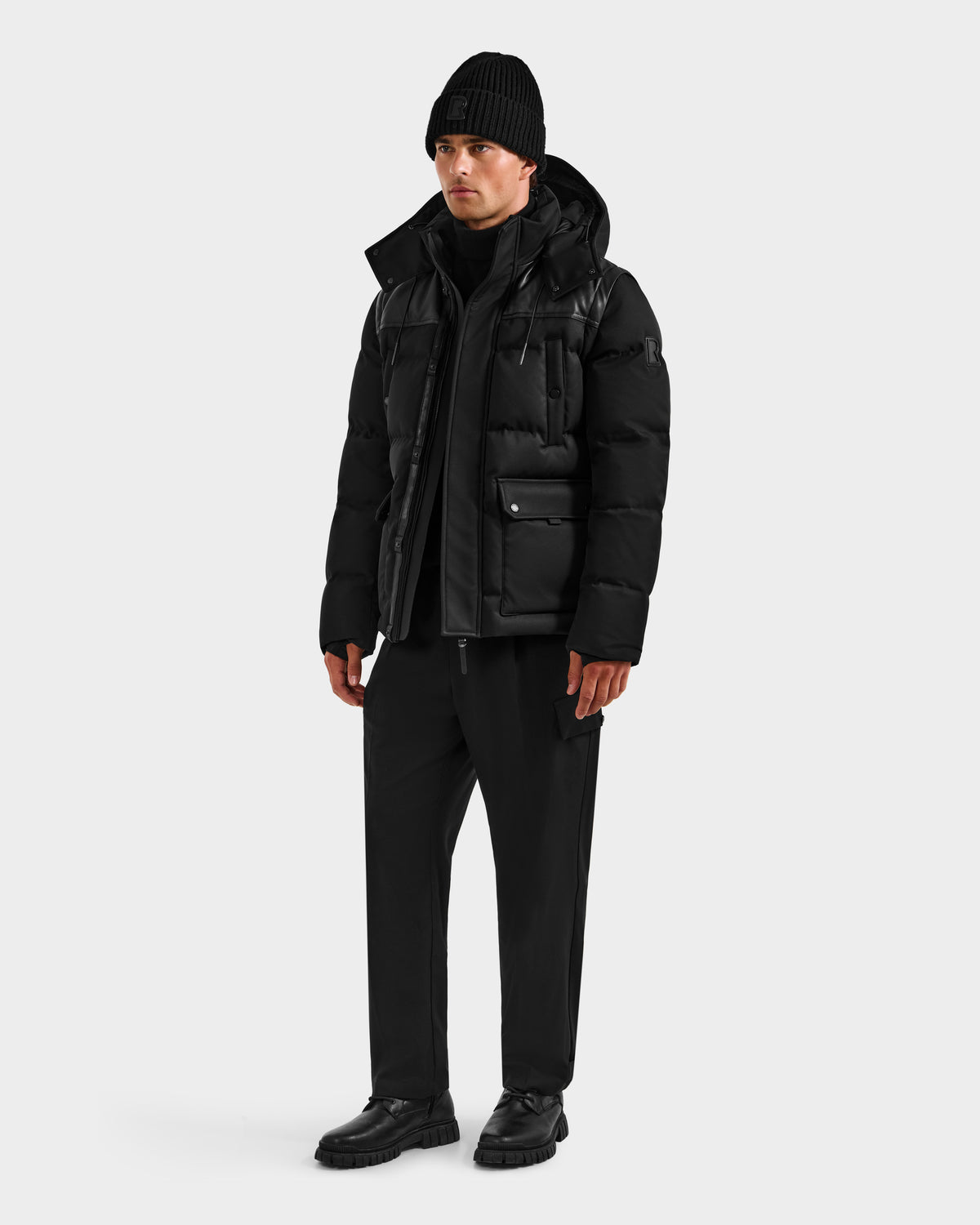 A man stands facing forward against a light gray background wearing an all-black winter look, featuring the Rudsak BALDWIN - 6124507 Black men's down parka, paired with a beanie, pants, and boots.