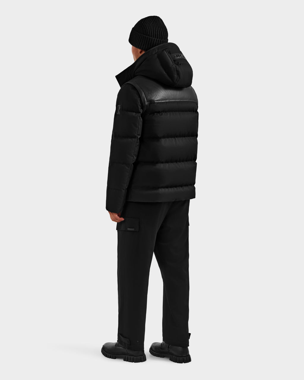 A person wearing the Rudsak BALDWIN - 6124507 Black men’s down parka with removable sleeves, a black beanie, black pants, and black shoes stands facing away from the camera against a plain light background.