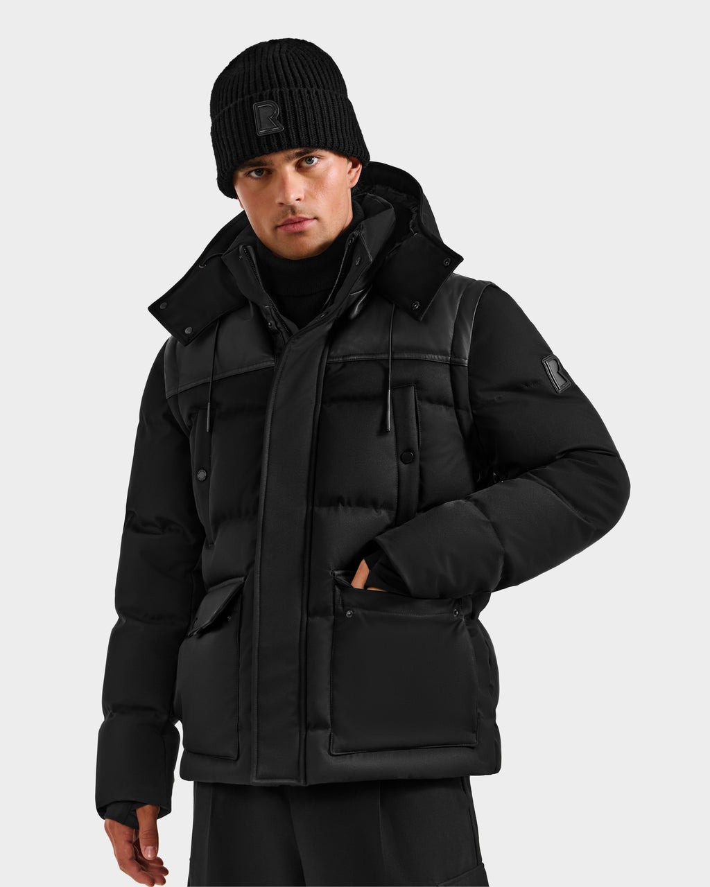 A man wears the Rudsak BALDWIN - 6124507 Black down parka with large pockets, a high collar, and removable sleeves, styled with a black beanie and pants against a plain light gray background.