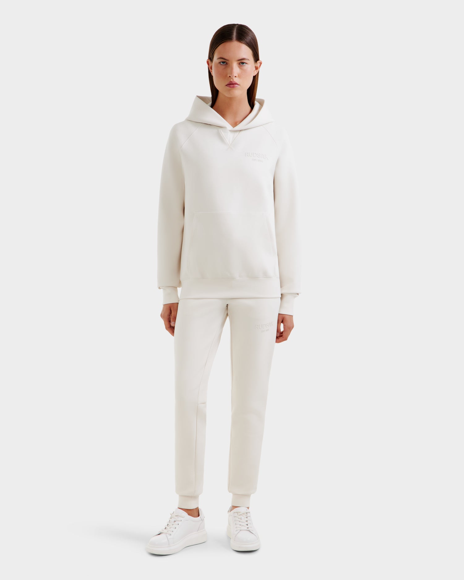 A person stands facing forward wearing the Rudsak BARRY - 9524011 Vanilla W unisex hoodie and matching jogger set with white sneakers against a plain white background.