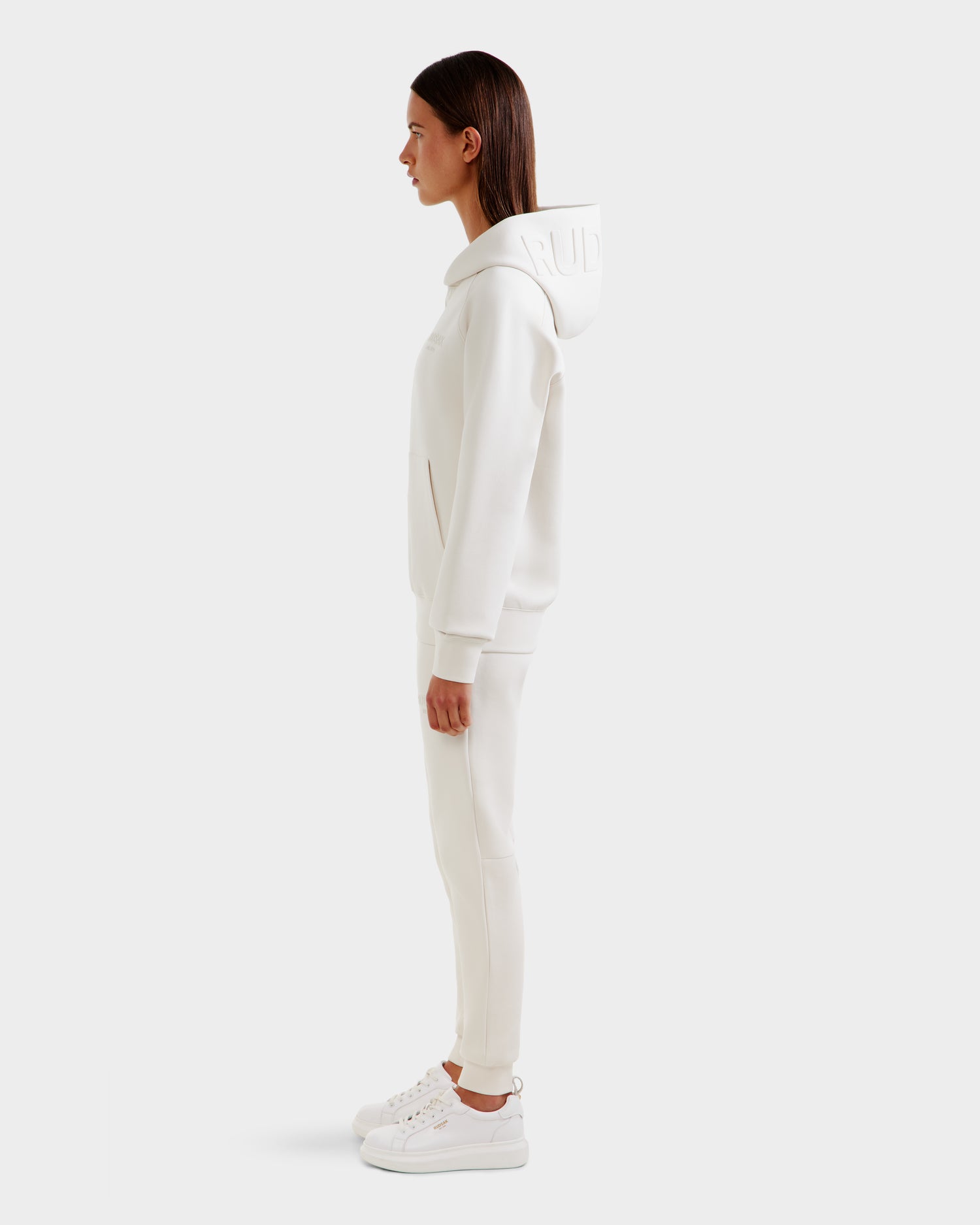 A woman stands in profile against a white background, wearing the Rudsak BARRY - 9524011 Vanilla W oversized hoodie with matching joggers and white sneakers. Her hair is straight and her hands are by her sides.