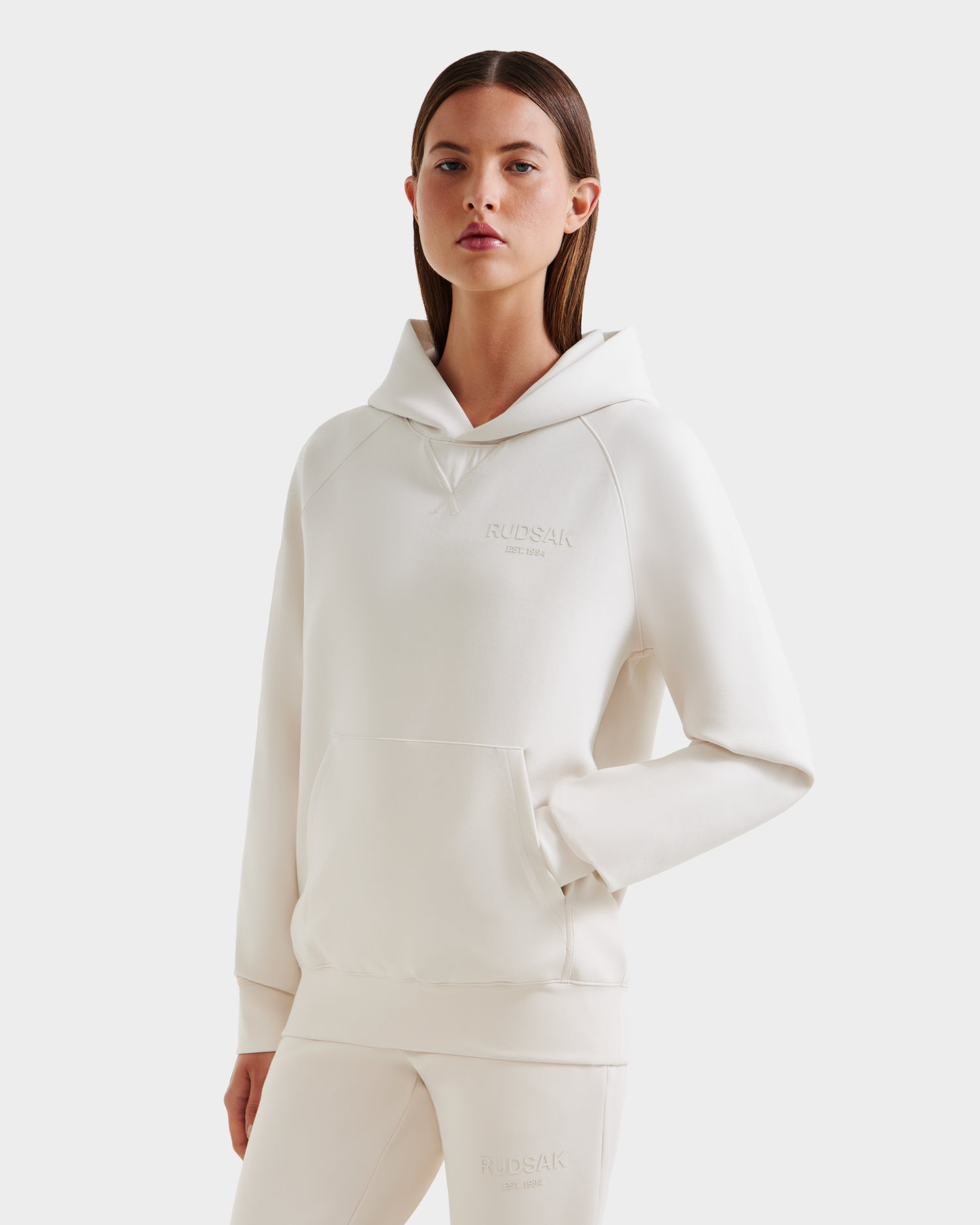 A woman with straight brown hair wears the Rudsak BARRY - 9524011 Vanilla W oversized hoodie and matching pants, standing with one hand in the sweatshirt pocket against a plain white background.