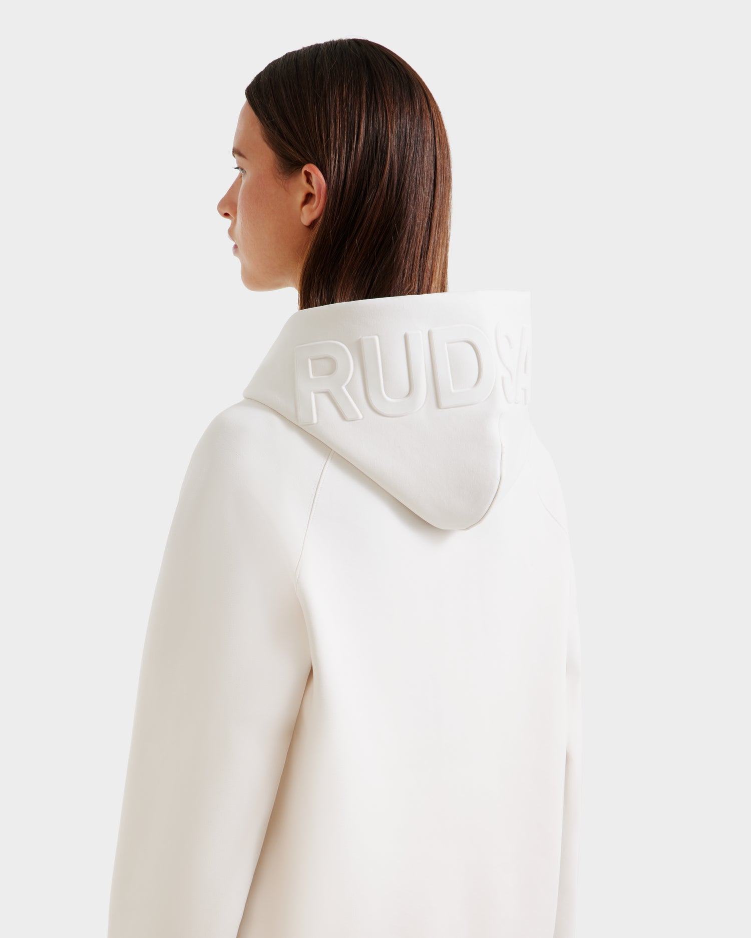 A person with straight brown hair wears the Rudsak BARRY - 9524011 Vanilla W coat with an oversized hoodie and embossed Rudsak lettering on the back, set against a plain, light-colored background.