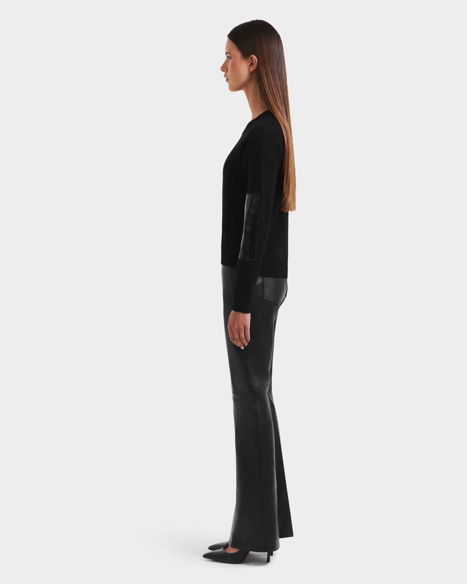 A woman stands in profile against a plain background, wearing the Rudsak BELLEROSE - 8525554 Black sweater with leather elbow patches, paired with black leather pants and pointed-toe shoes. Her straight brown hair is worn down.