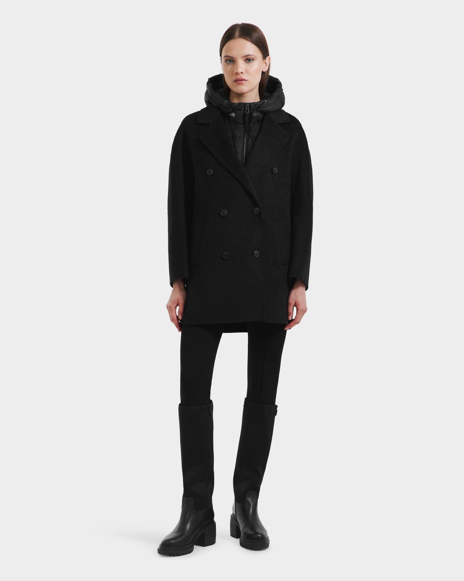 A woman faces forward wearing the Rudsak BETHANY - 8125625 Black wool-silk-yak modular coat, paired with black pants, knee-high black boots, and a black hooded inner layer against a plain white background.